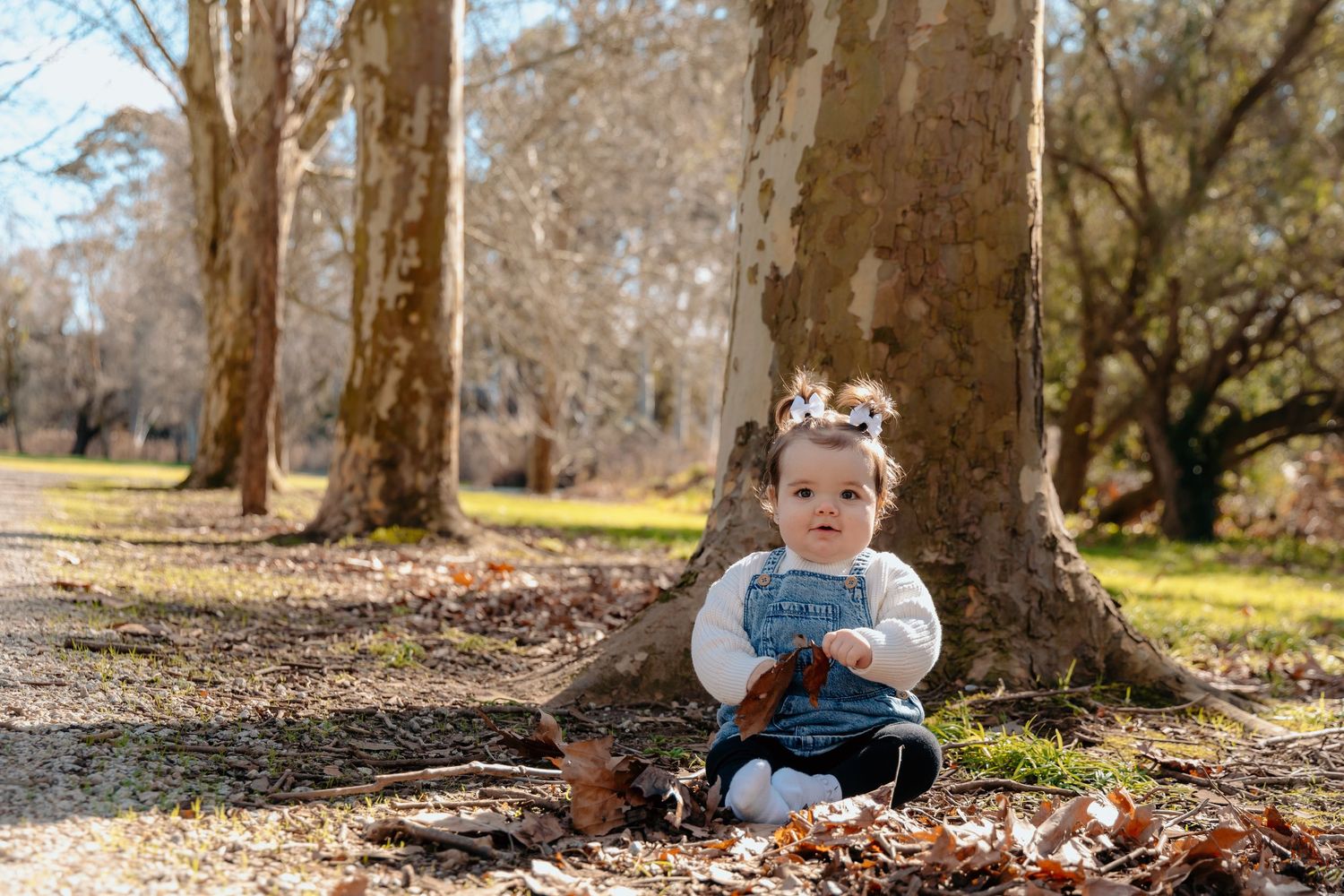 A small child sits on autumn leaves in a sunny park wearing a blue outfit and floral headband.