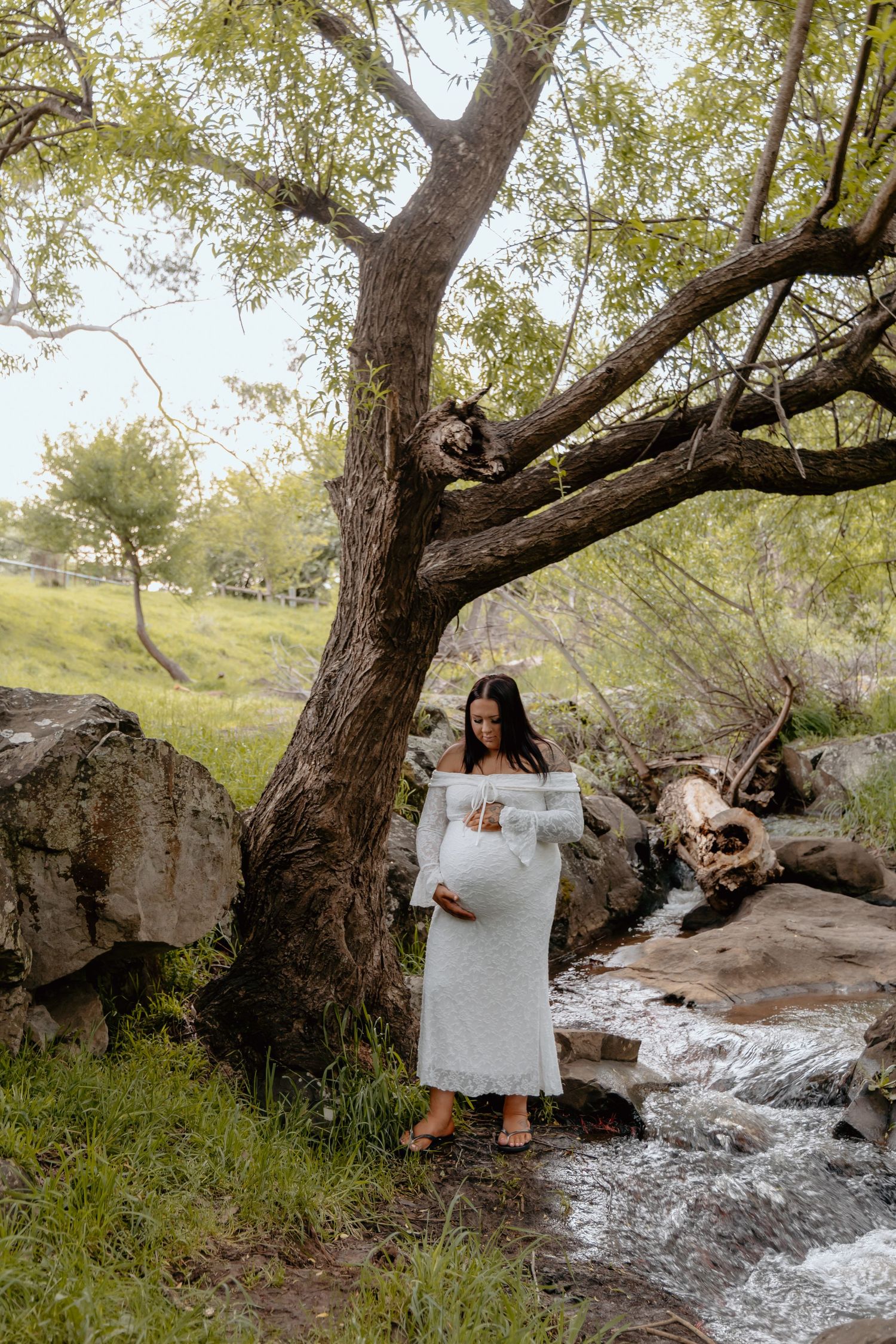 A person in white dress sits beside a flowing stream under a large tree in a natural outdoor setting.