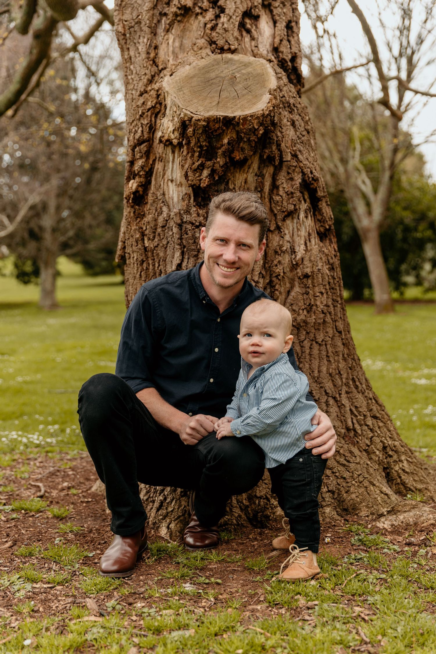 A tender outdoor portrait by a large tree trunk in a green park setting.