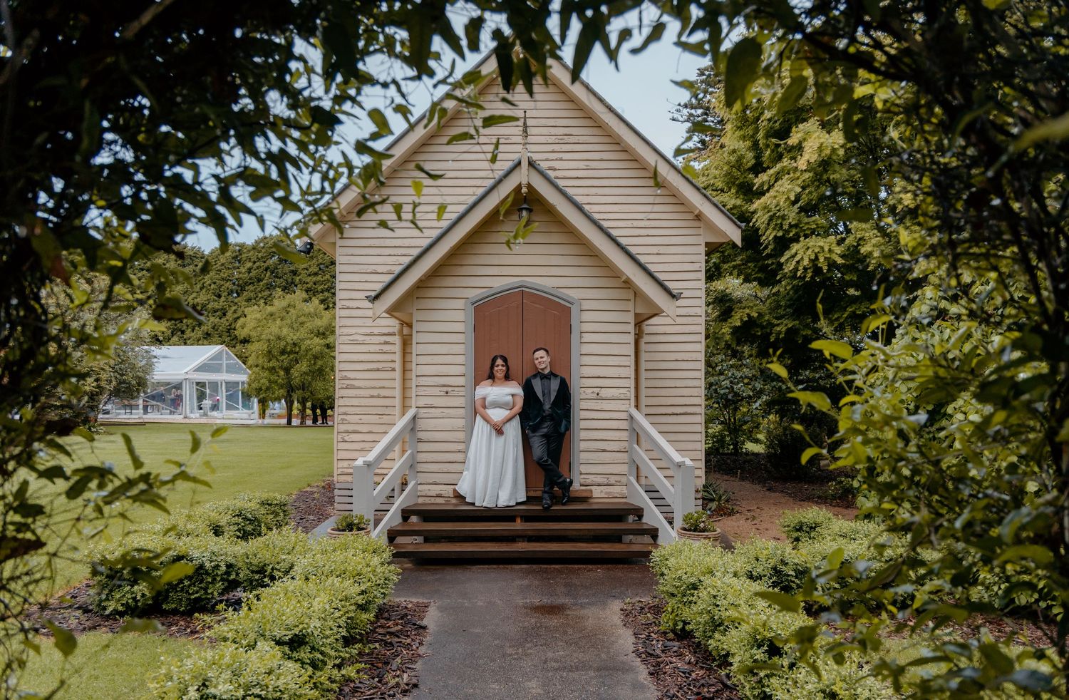 A rustic white church building with red doors sits nestled among green trees on a sunny day.