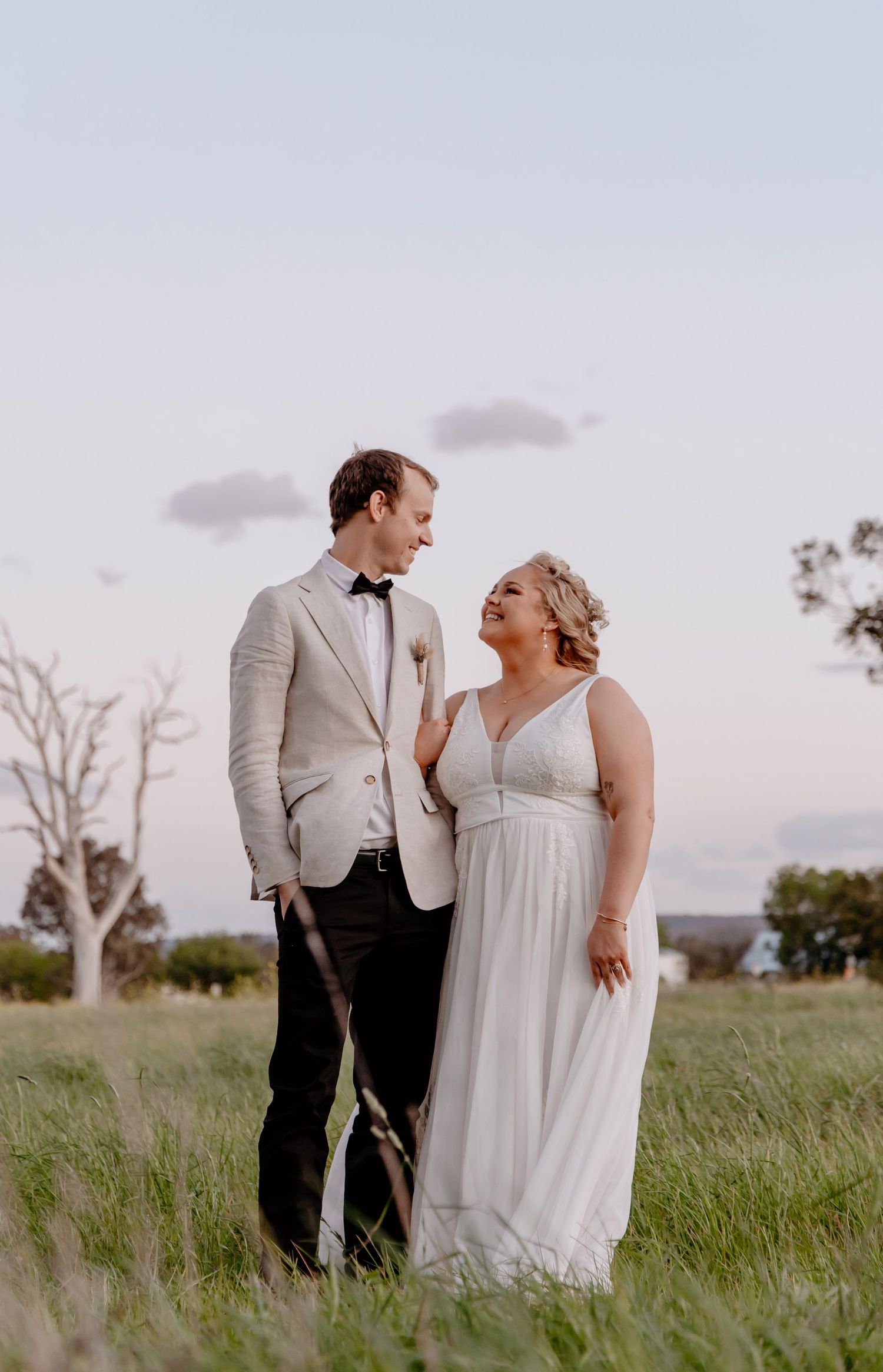 A romantic couple in wedding attire stands together in a grassy field at sunset, sharing intimate moments.