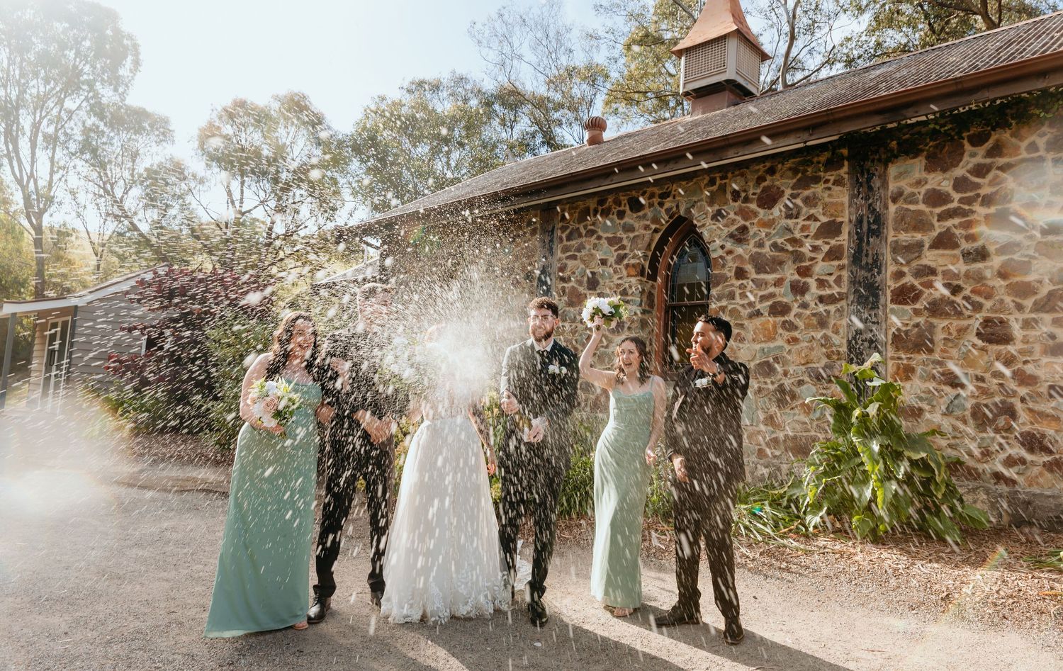A wedding party poses together in sage green dresses against a rustic stone wall on a sunny afternoon.