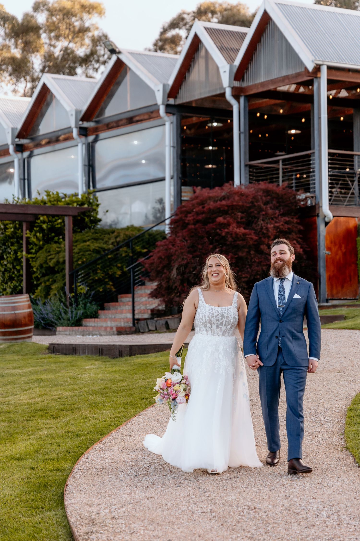 Couple walks along gravel path beside white modern chapel buildings with string lights and landscaped grounds.