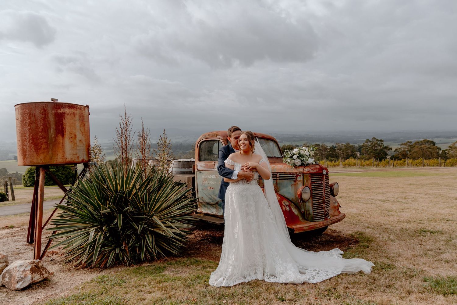 Romantic couple in wedding attire poses beside rustic vintage truck and desert plants on cloudy day.
