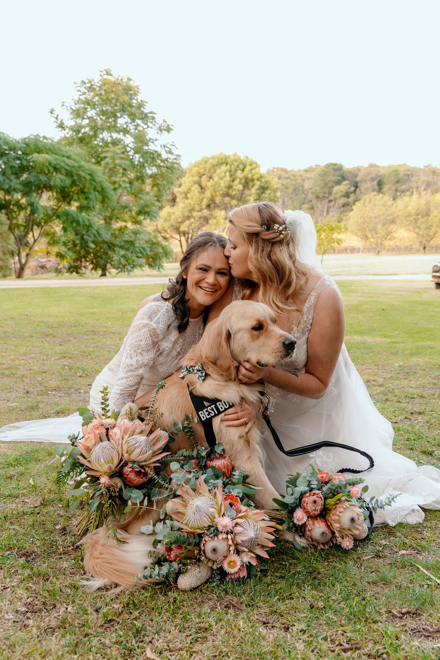 Two people in wedding attire sit on grass with a golden retriever and pink floral bouquets at an outdoor celebration.