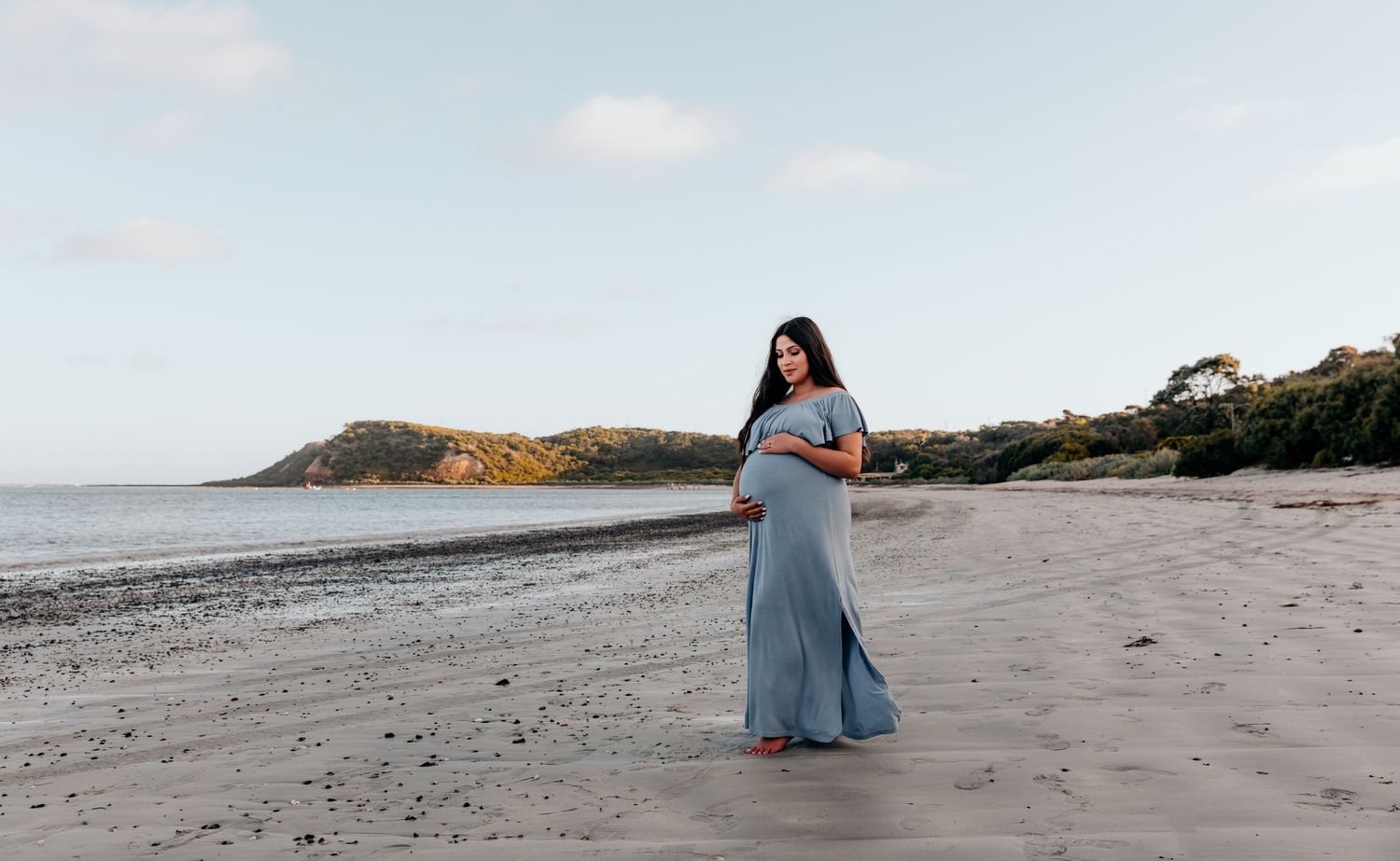 Pregnant woman in blue dress standing alone on beach at sunset.