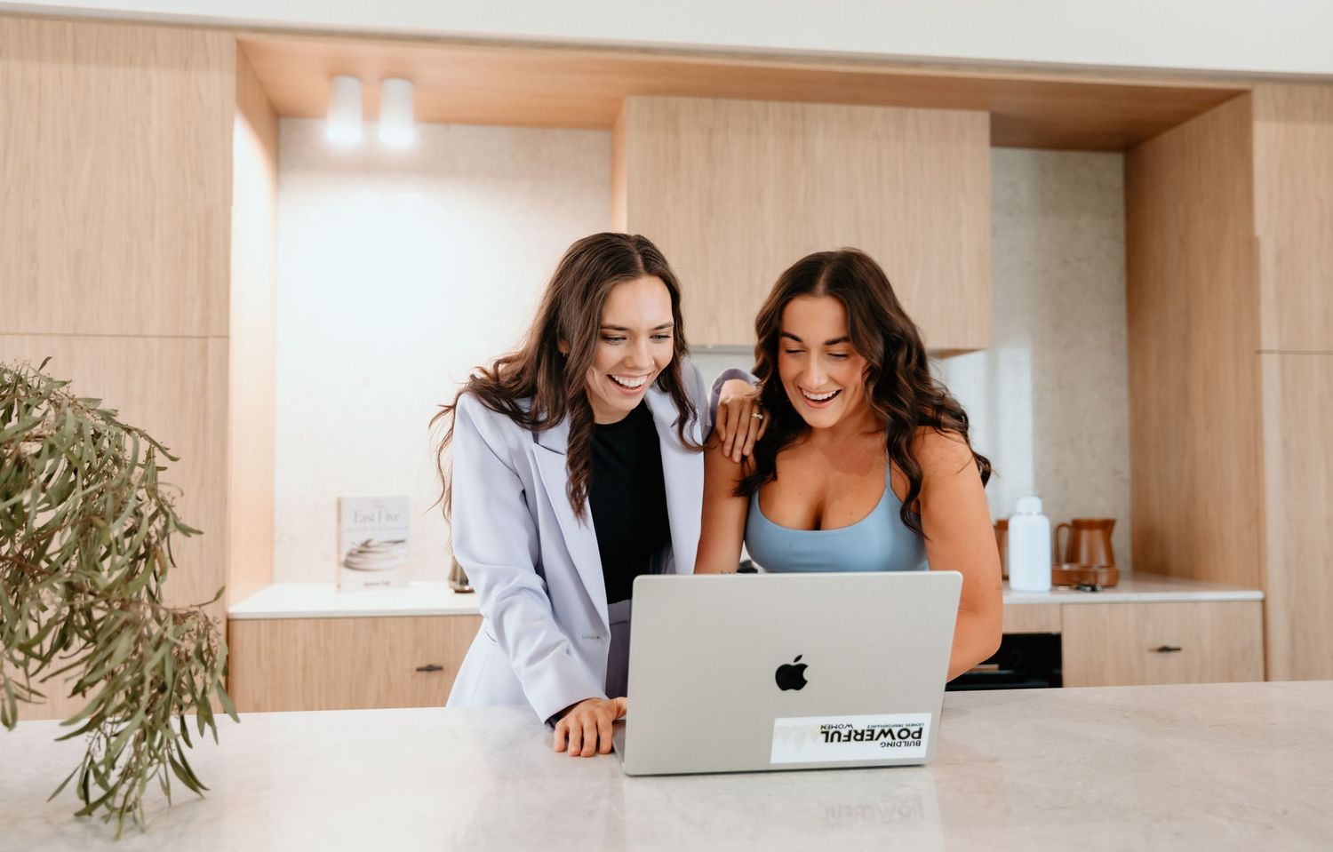Two women looking at laptop together in modern kitchen with wood cabinets.