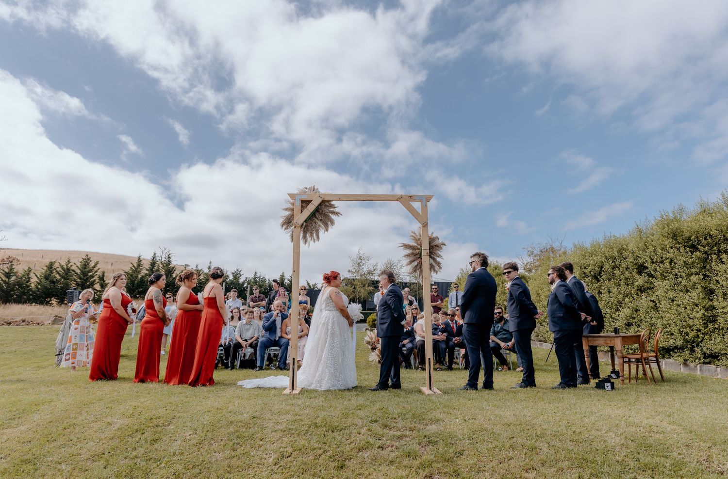 Outdoor wedding ceremony on green lawn with wooden arch, red bridesmaid dresses, and guests seated under blue sky.