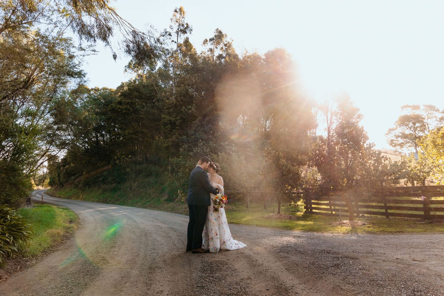 Bride and groom share a romantic kiss on a sunlit gravel path surrounded by lush trees and a wooden fence.