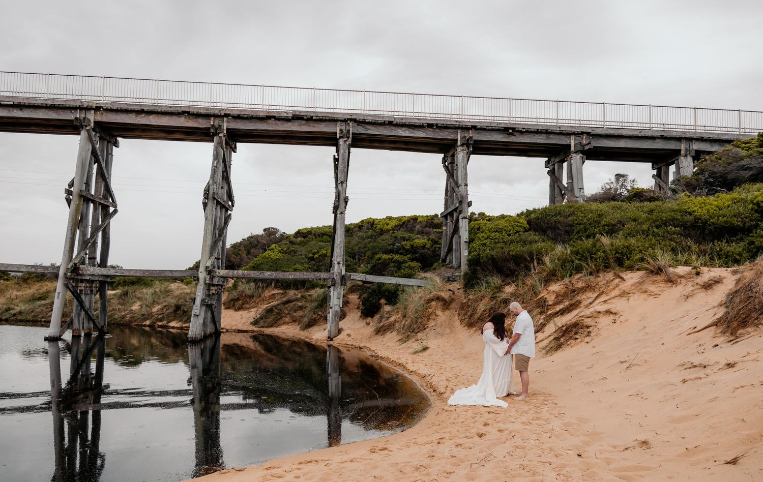 A couple in wedding attire poses beneath a wooden bridge on a sandy beach.