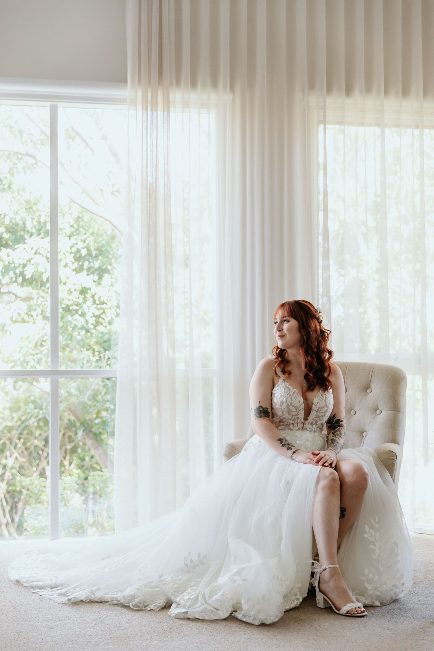 Elegant bride in flowing white lace gown lounges gracefully in tufted chair by sunlit window with sheer curtains.