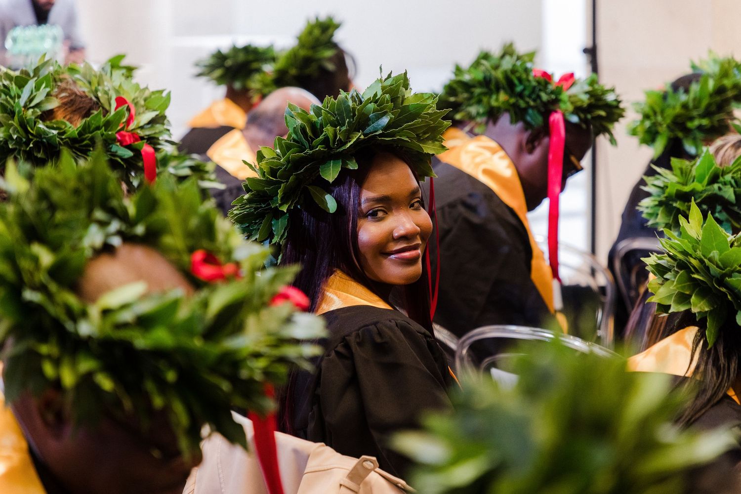 Smiling graduate in black gown wearing green leaf crown at graduation ceremony with fellow graduates in floral wreaths.