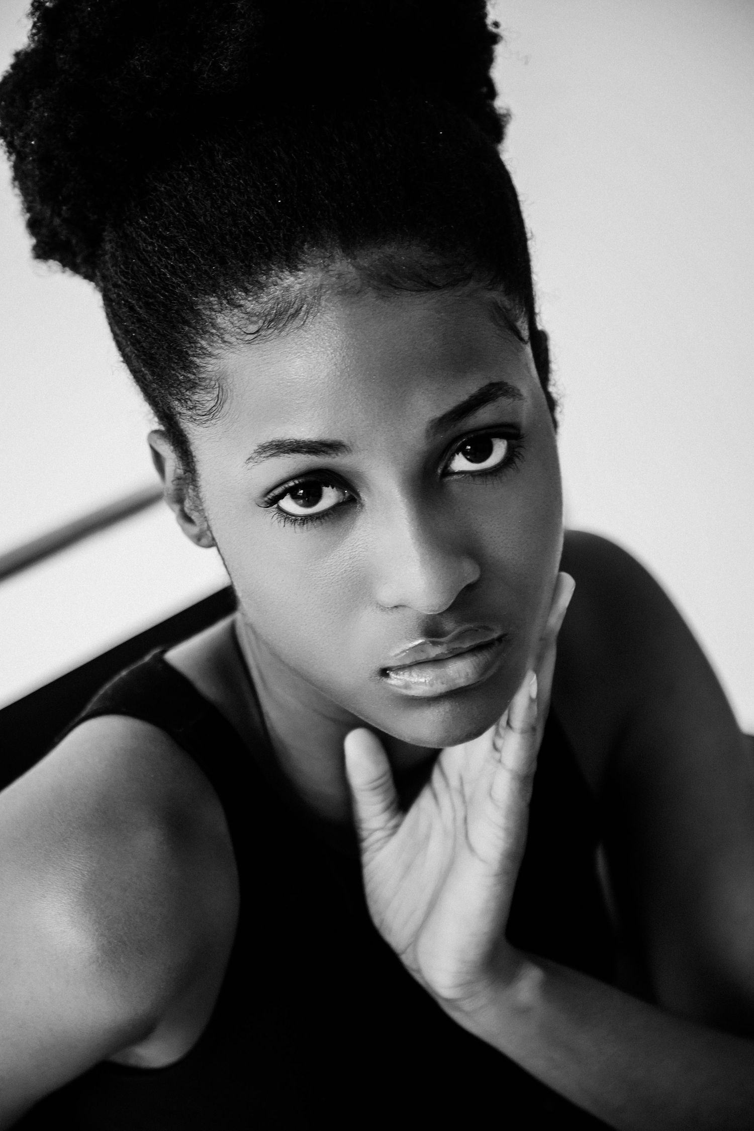 Black and white portrait of a young woman with natural updo hairstyle, resting chin on hand, wearing black top.