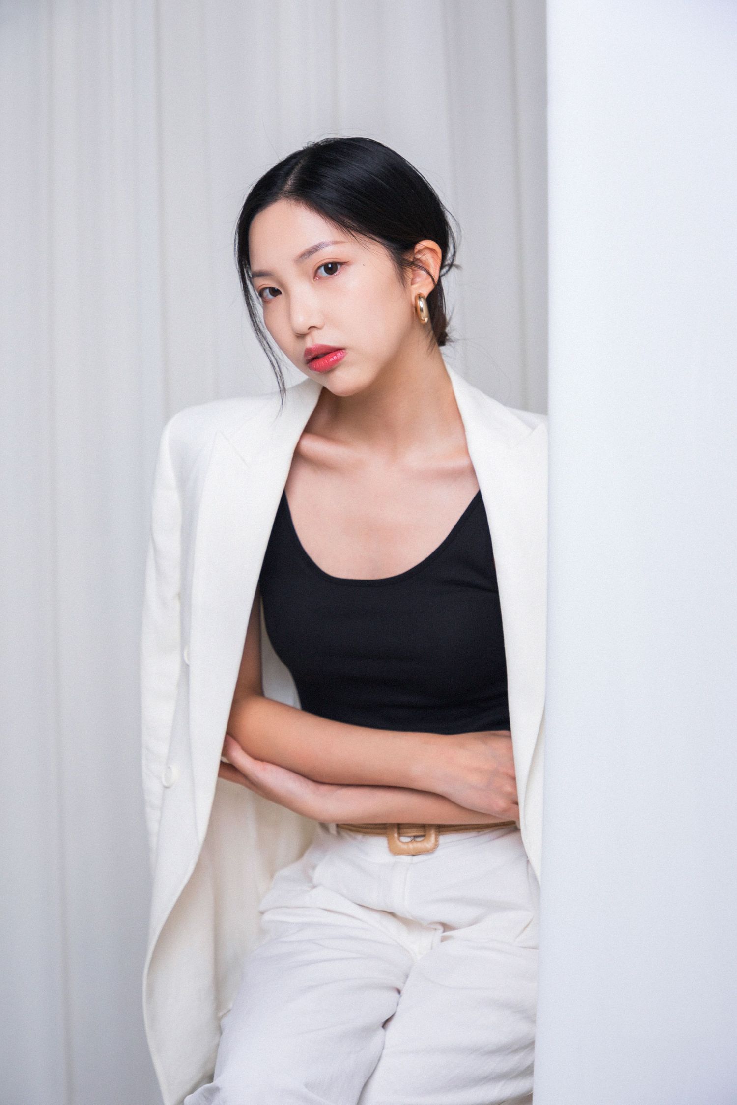 Young Asian woman in white blazer and black top posing confidently with arms crossed against a white wall.