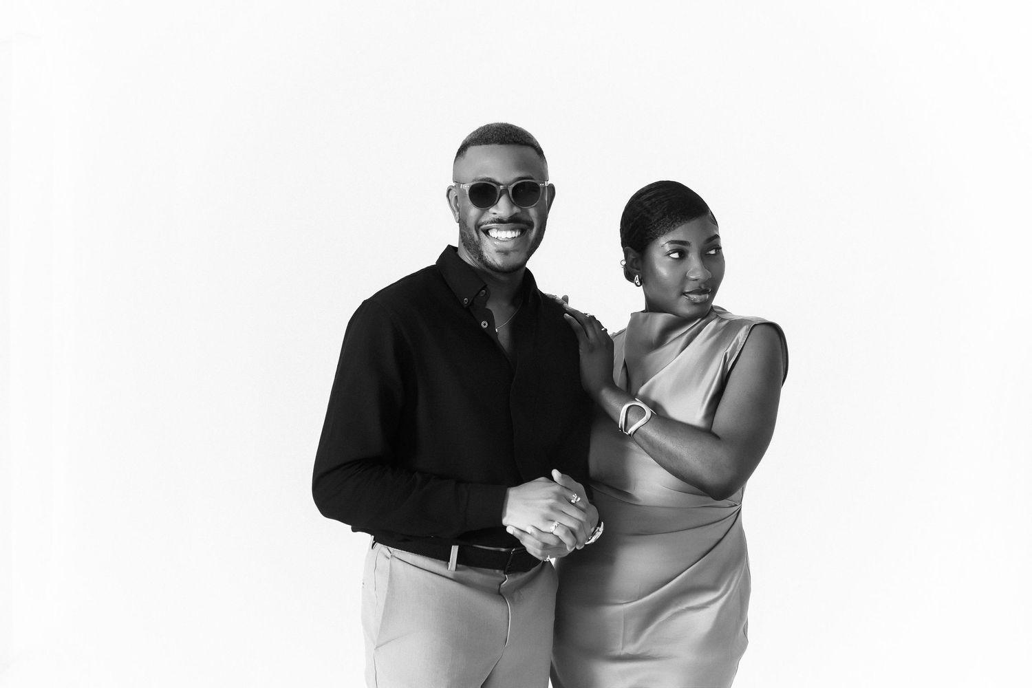 Black and white portrait of a smiling couple posing together, man in sunglasses and black shirt, woman in elegant dress.