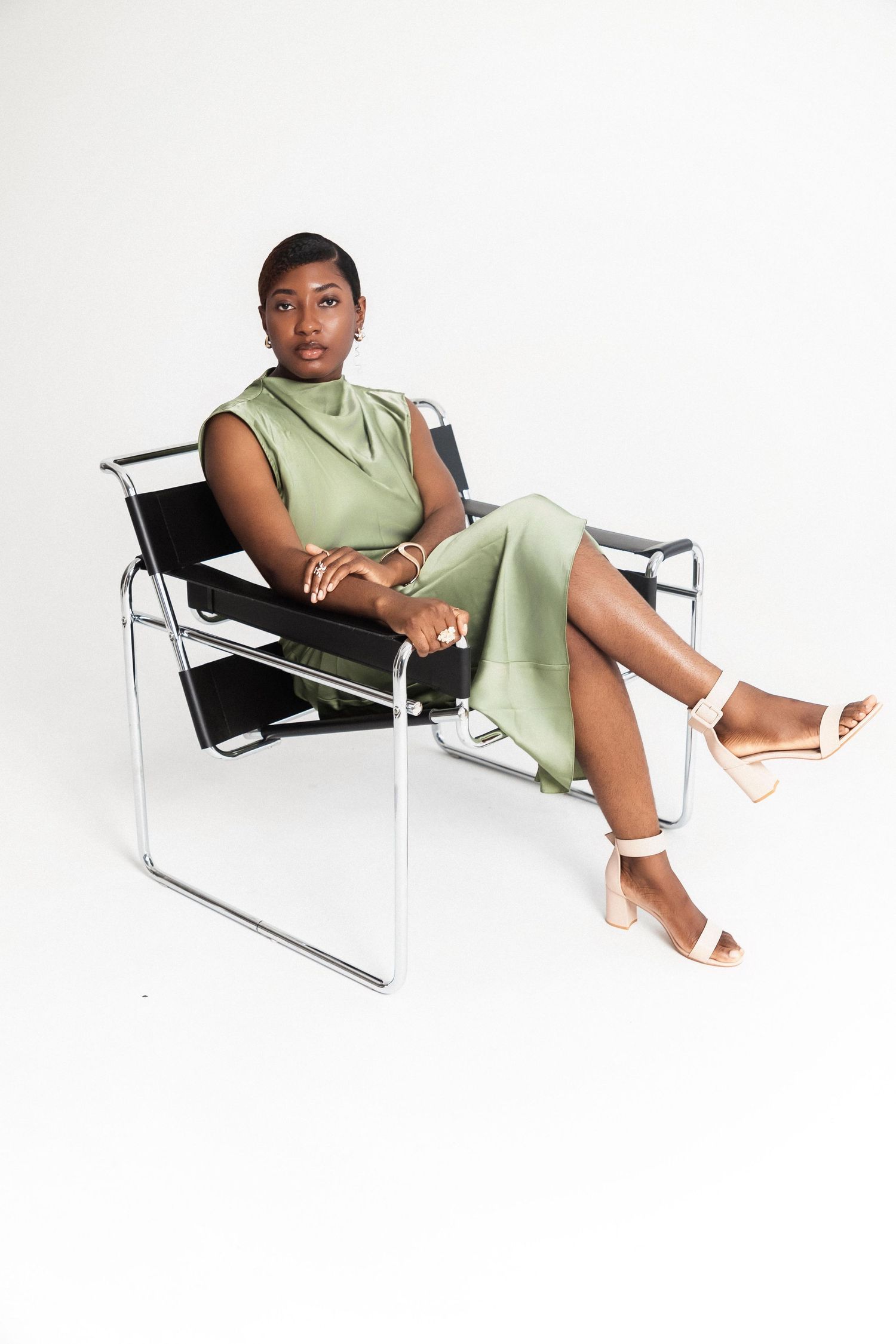 Woman in sage green dress and white heeled sandals sitting in a modern Wassily chair against white background.