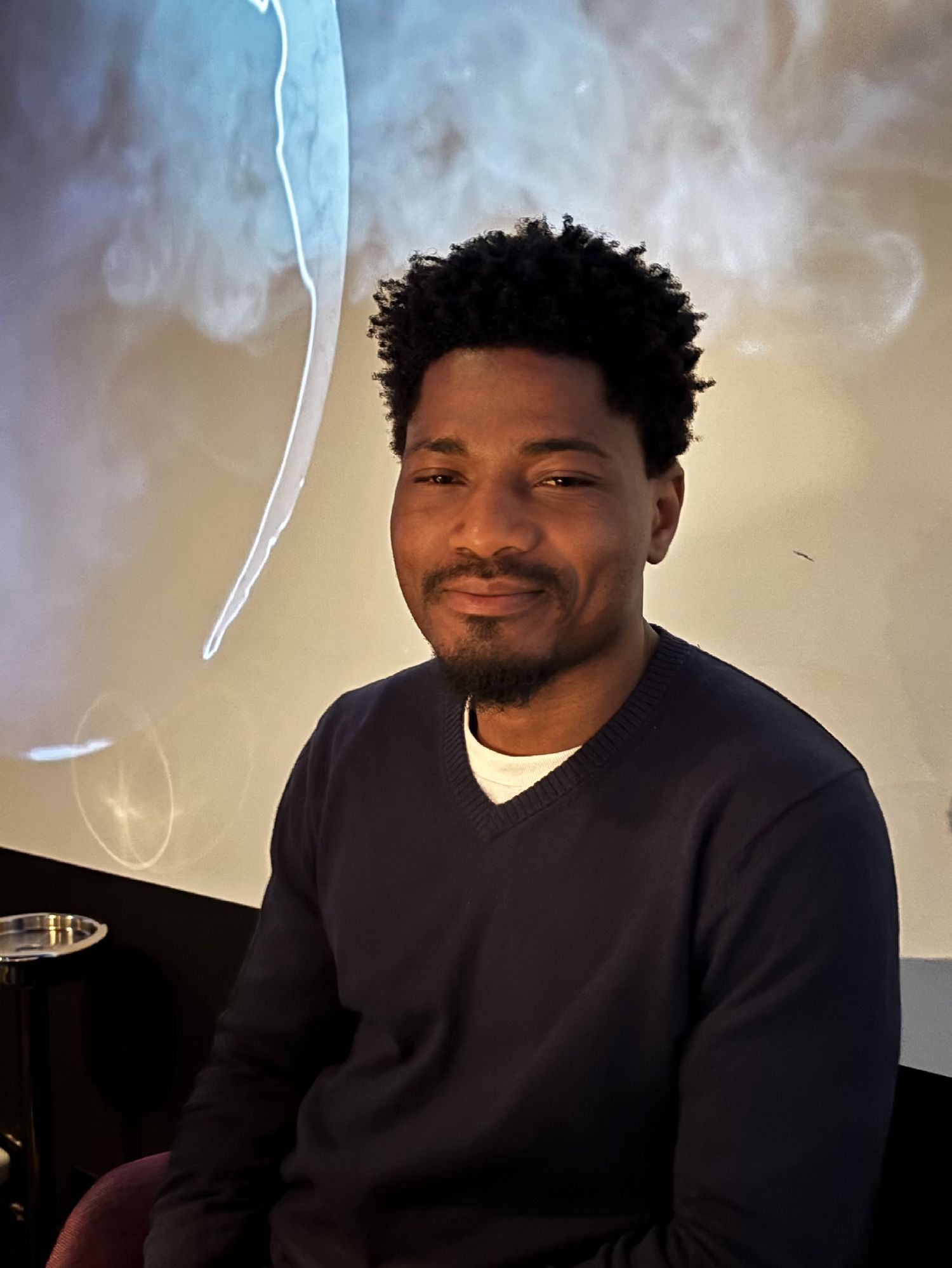 Young man with short curly hair and beard wearing a navy sweater, smiling in a dimly lit room with smoky background.
