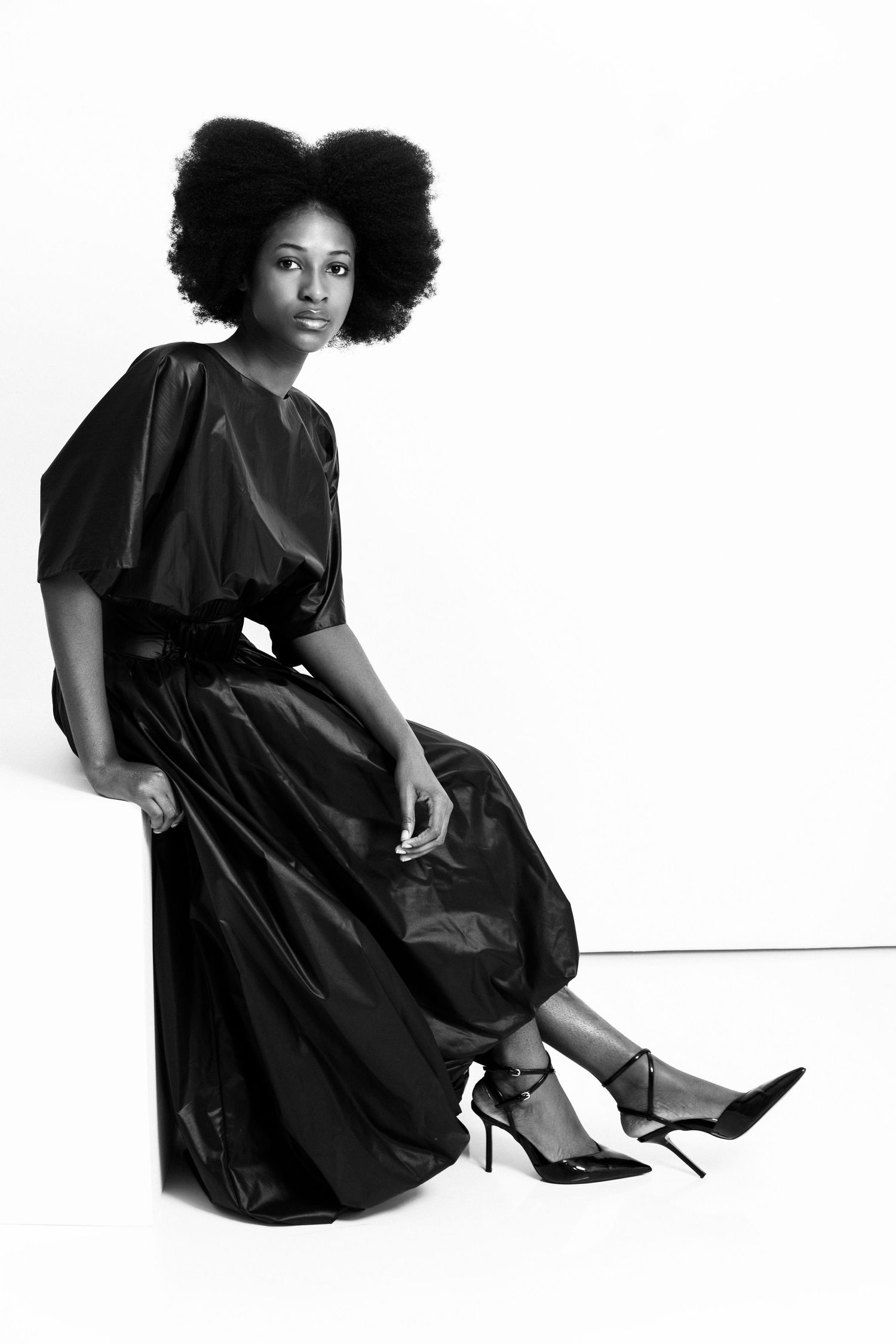 Black and white photo of a woman in an elegant black satin dress with afro hair, seated gracefully in heels.