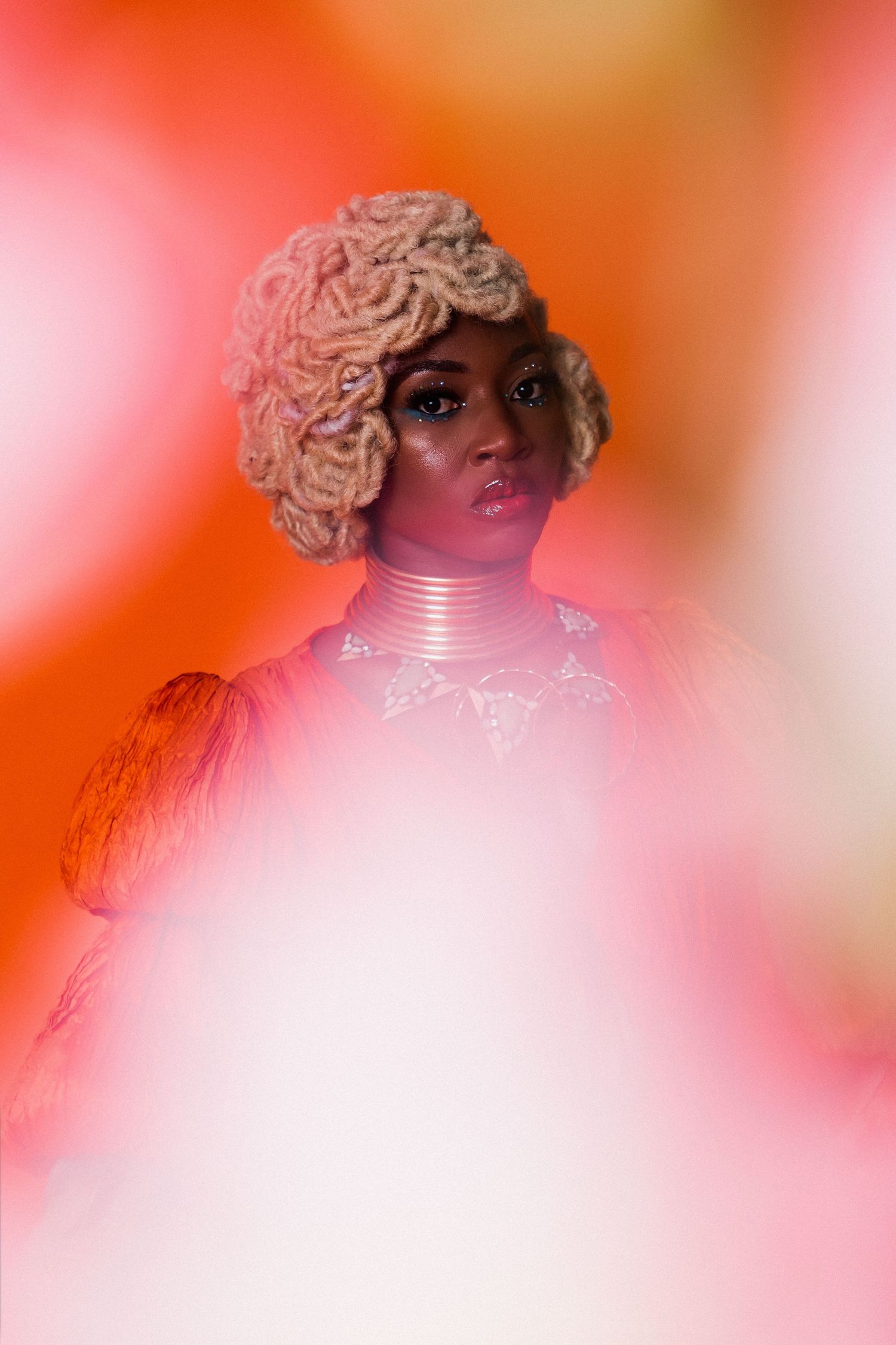 A Black woman in a pink yarn wig and neck rings poses against a vibrant orange-pink background.