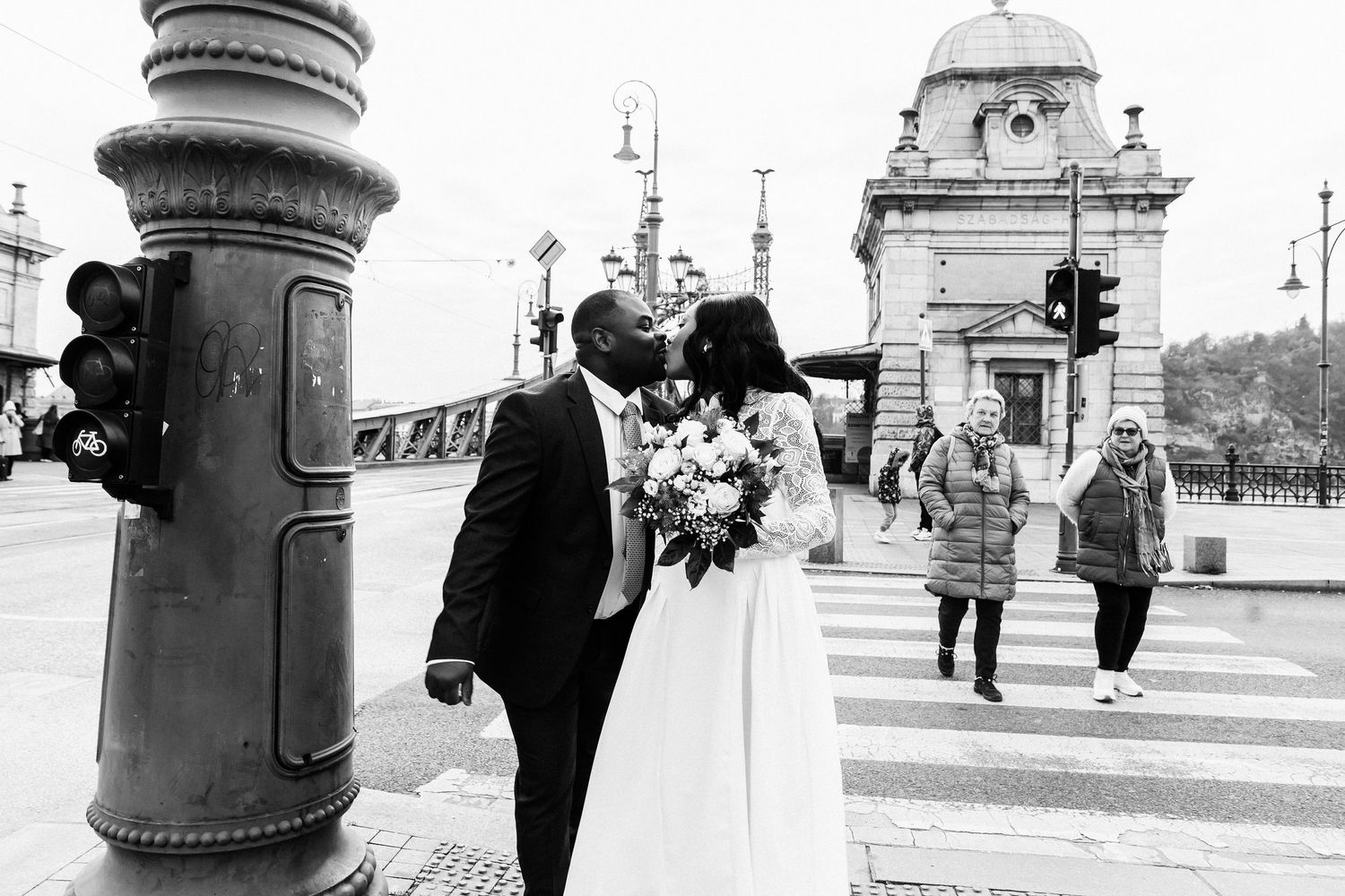 Black and white photo of a couple kissing on a city street, bride holding bouquet, pedestrians crossing in background.