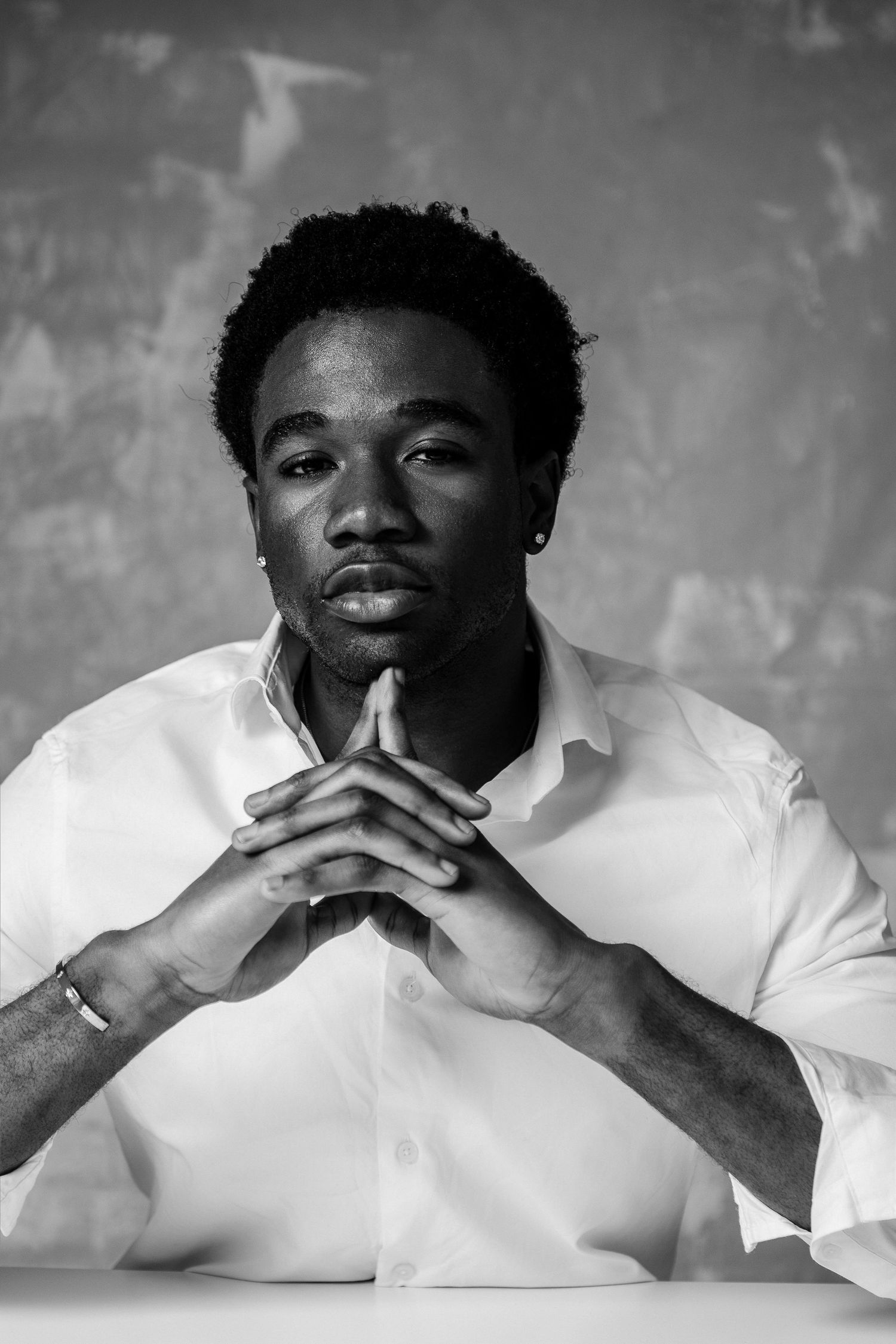 Black and white portrait of a young man in white shirt with hands clasped, looking confidently at camera.