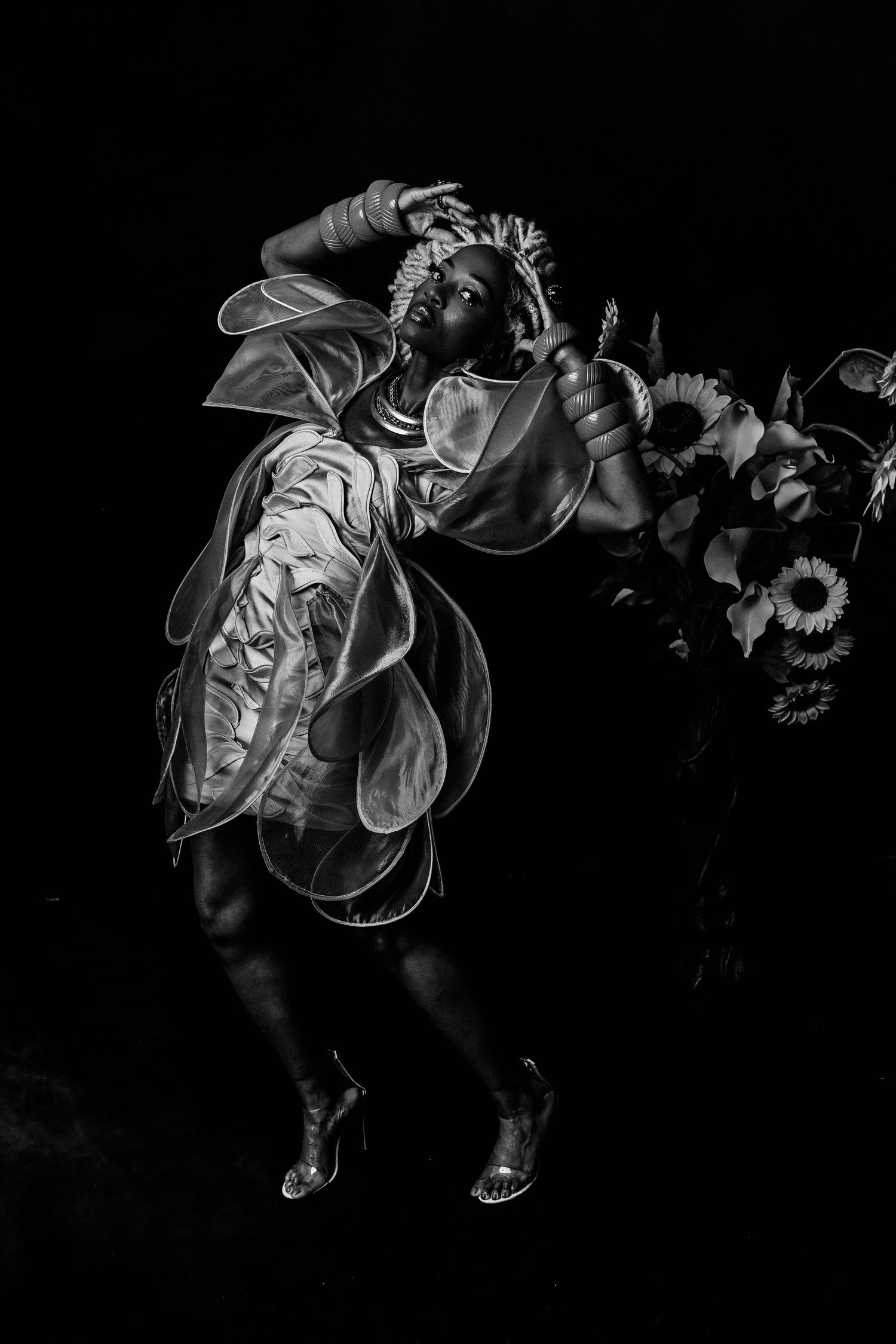 Black and white photo of a performer in an elaborate floral costume holding sunflowers against a dark background.