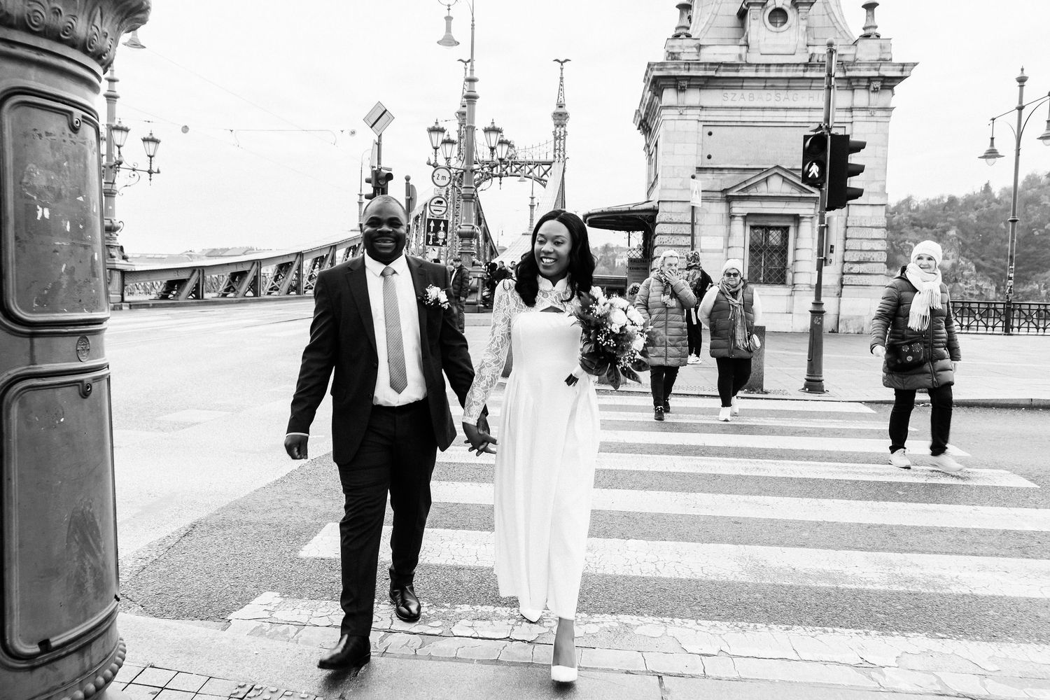 Black and white photo of a couple in wedding attire holding hands at a crosswalk in an urban European city.