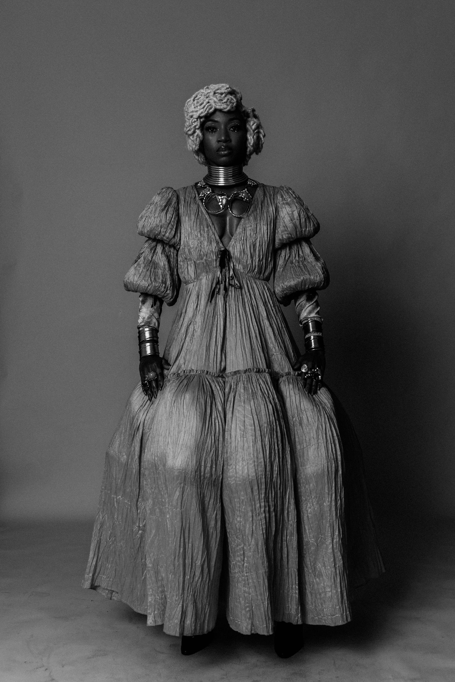Black and white portrait of a woman in a flowing tiered dress with puff sleeves, ornate jewelry, and floral headpiece.