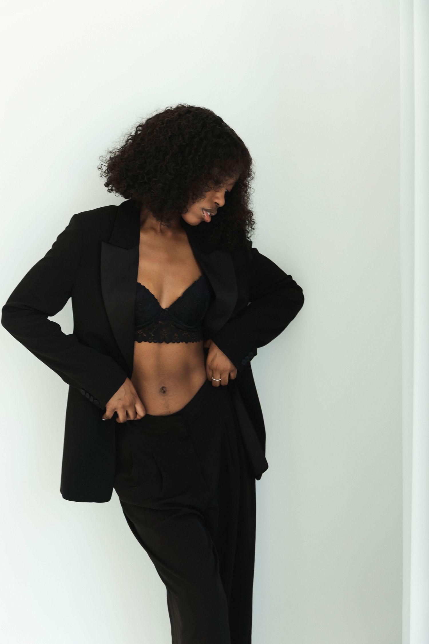 Woman in black blazer and lace bralette posing confidently against white wall with curly hair.