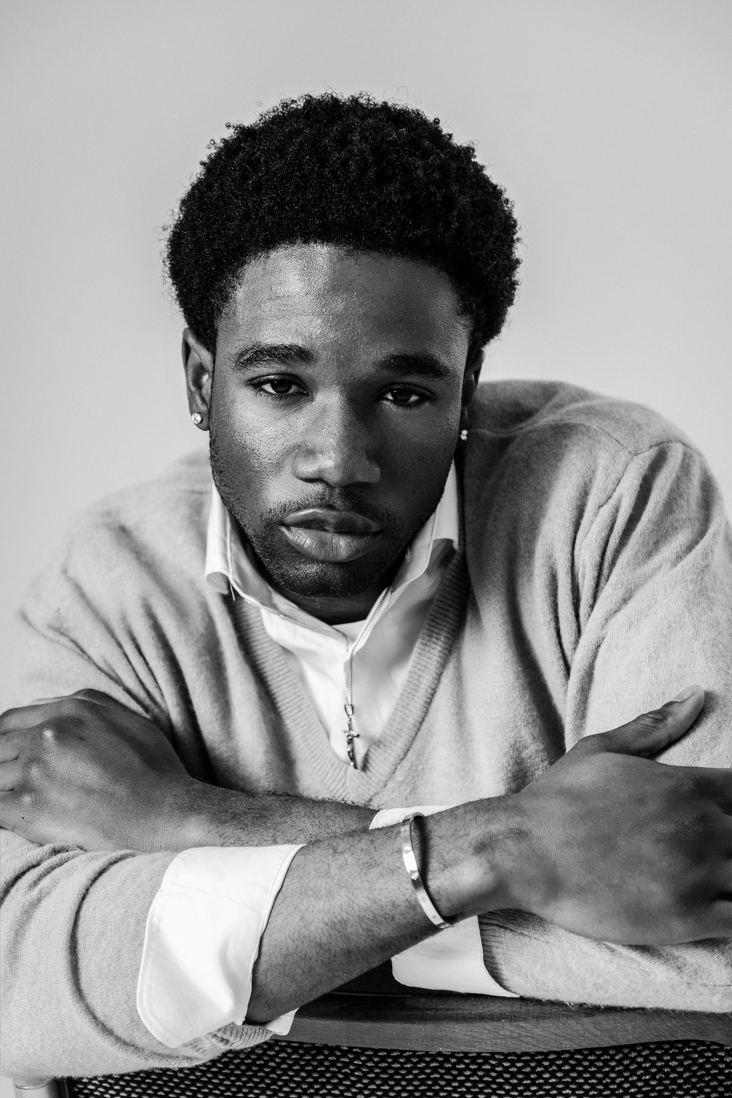 Black and white portrait of a young man in a sweater leaning forward with arms crossed, wearing a cross necklace.