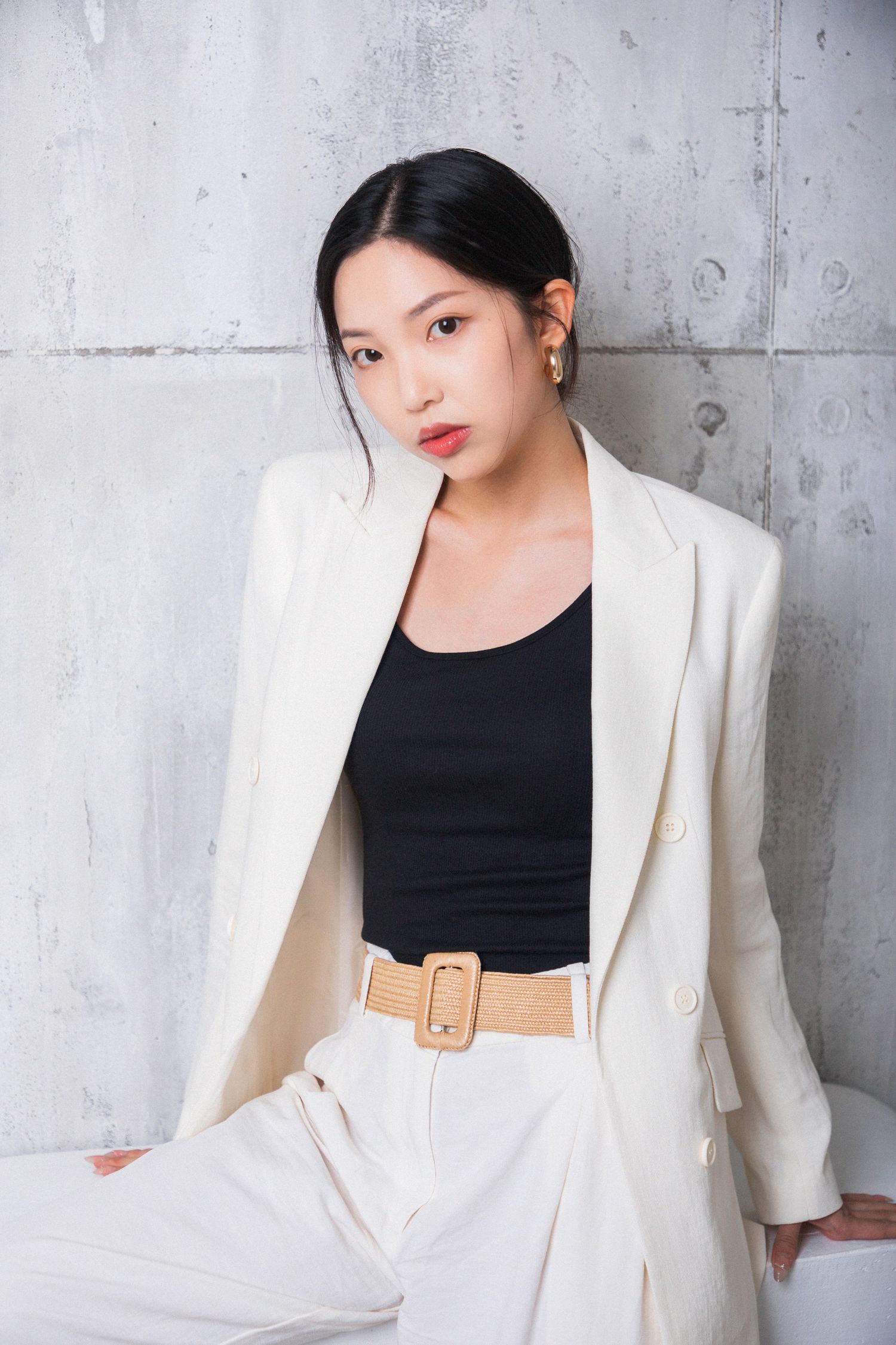 Young Asian woman in white blazer and pants with black top and woven belt poses against concrete wall.