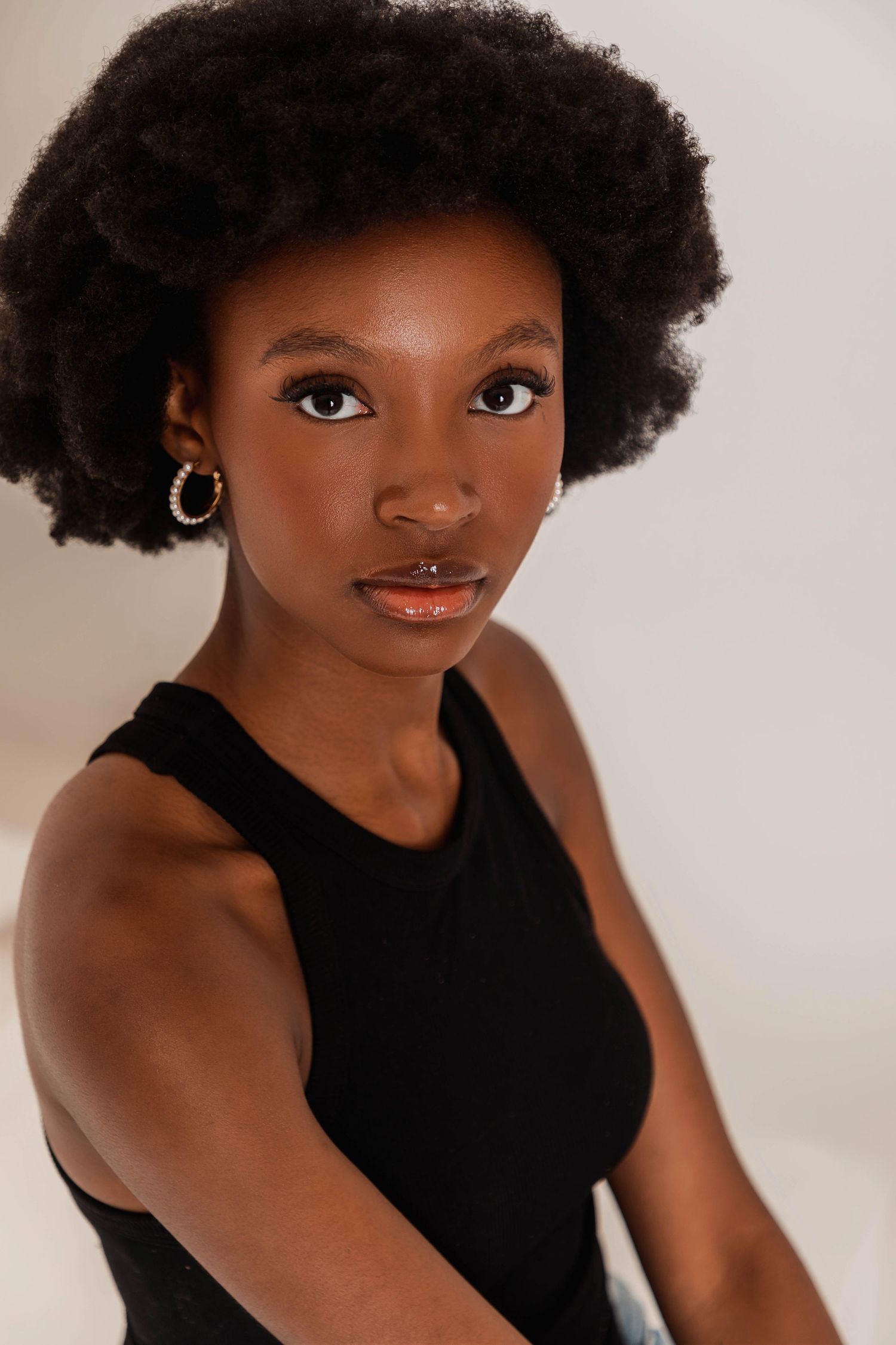 Young Black woman with natural afro hair, hoop earrings, nose ring, wearing black sleeveless top, posing against light background.