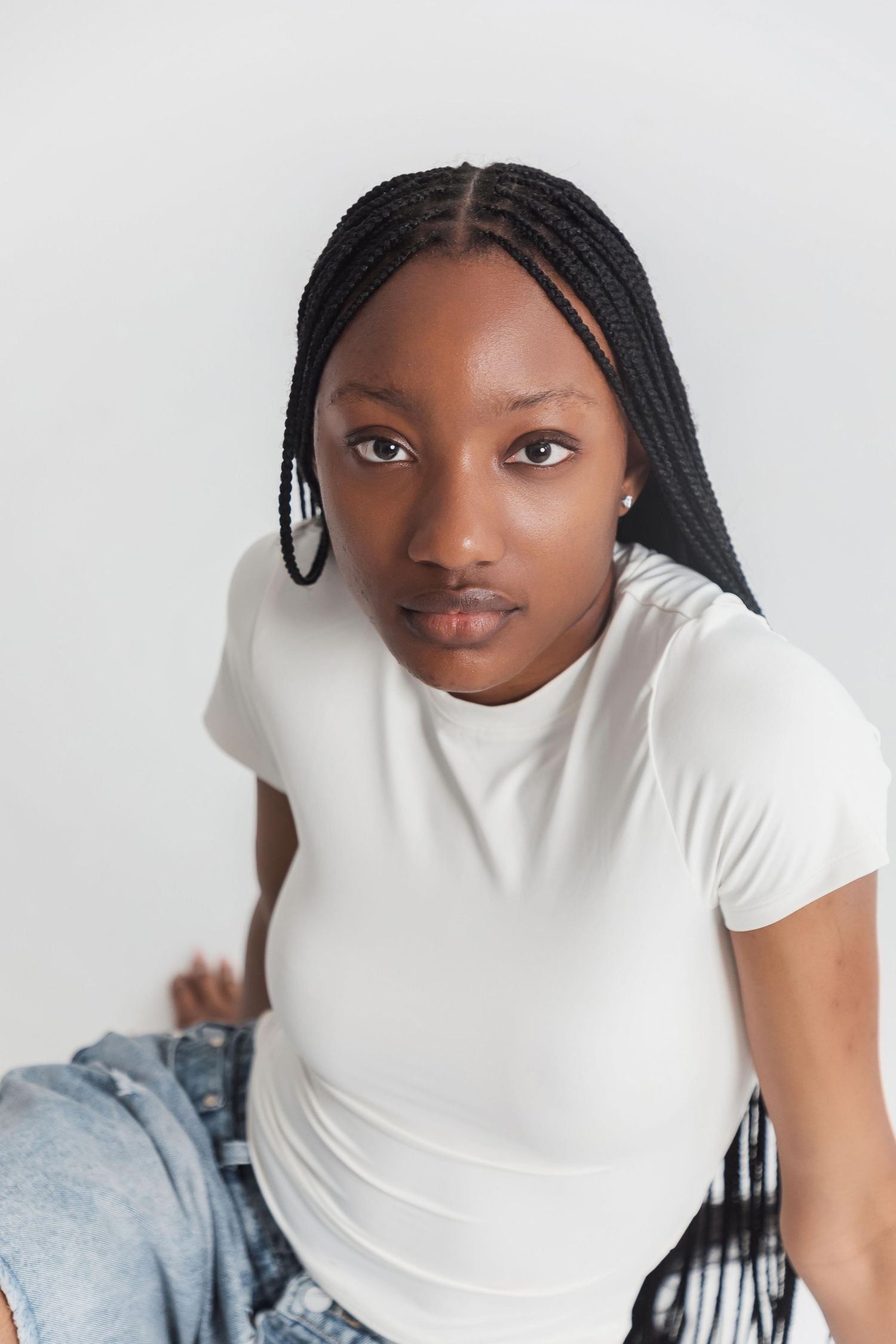 Young Black woman with box braids wearing a white t-shirt and jeans, sitting against a light gray background.