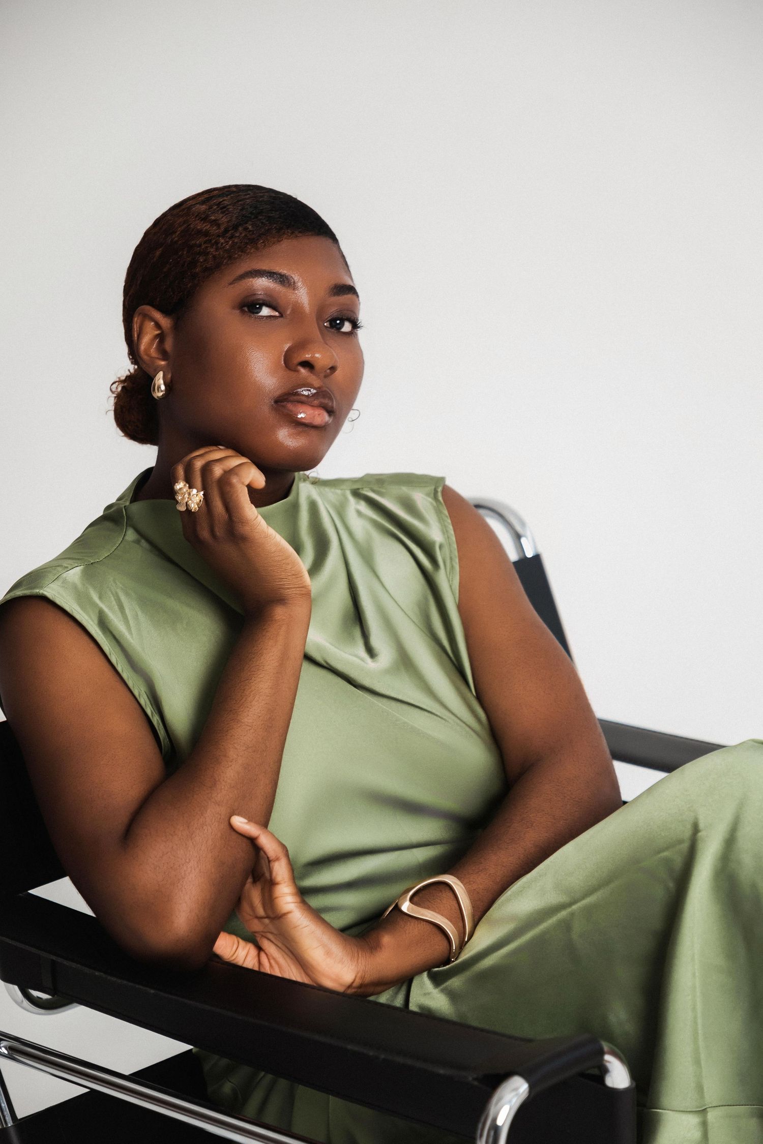 Woman in green satin outfit seated in black chair, wearing gold jewelry, posing thoughtfully against white background.