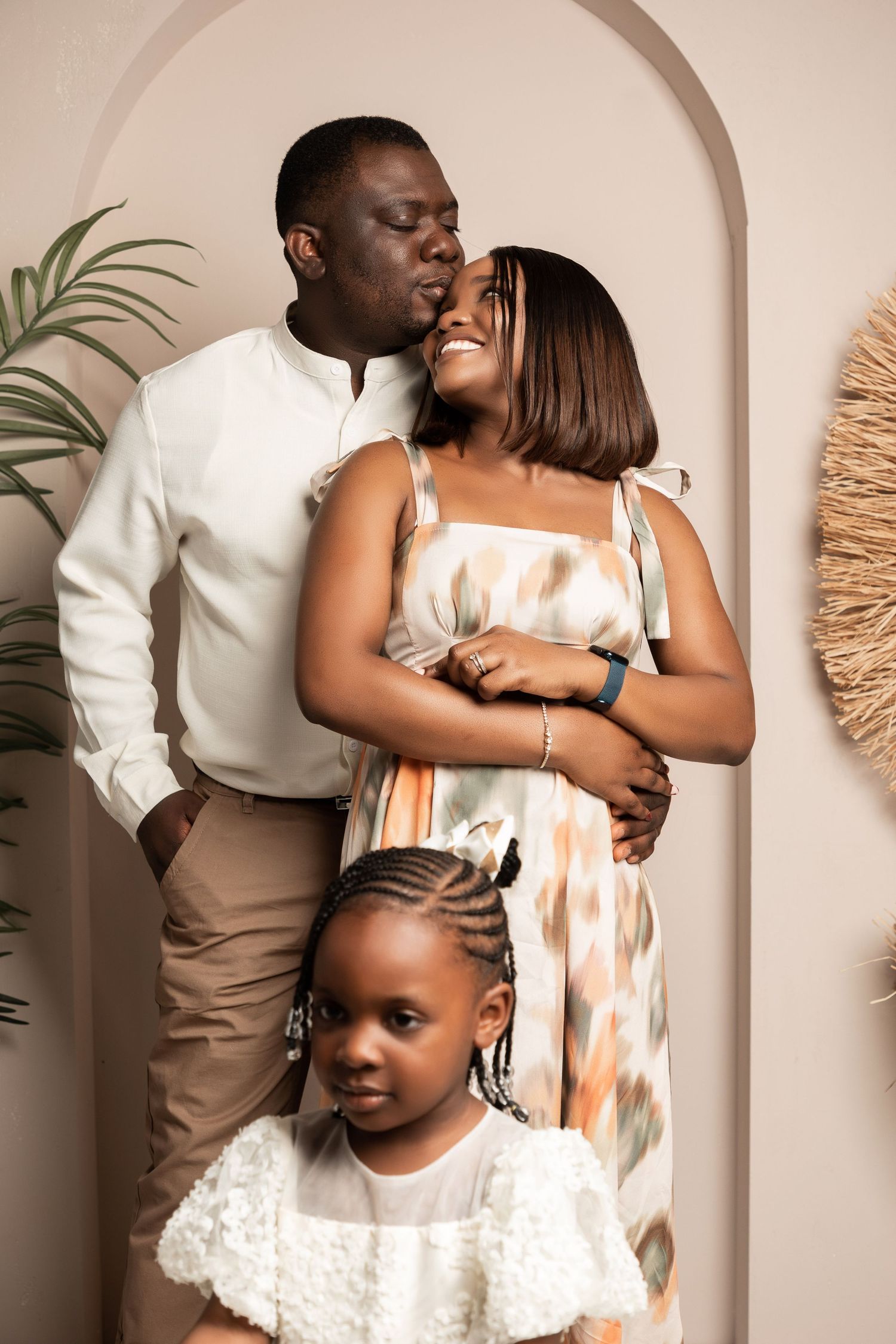 A loving Black family portrait with a man kissing a woman's forehead while their young daughter stands in front.