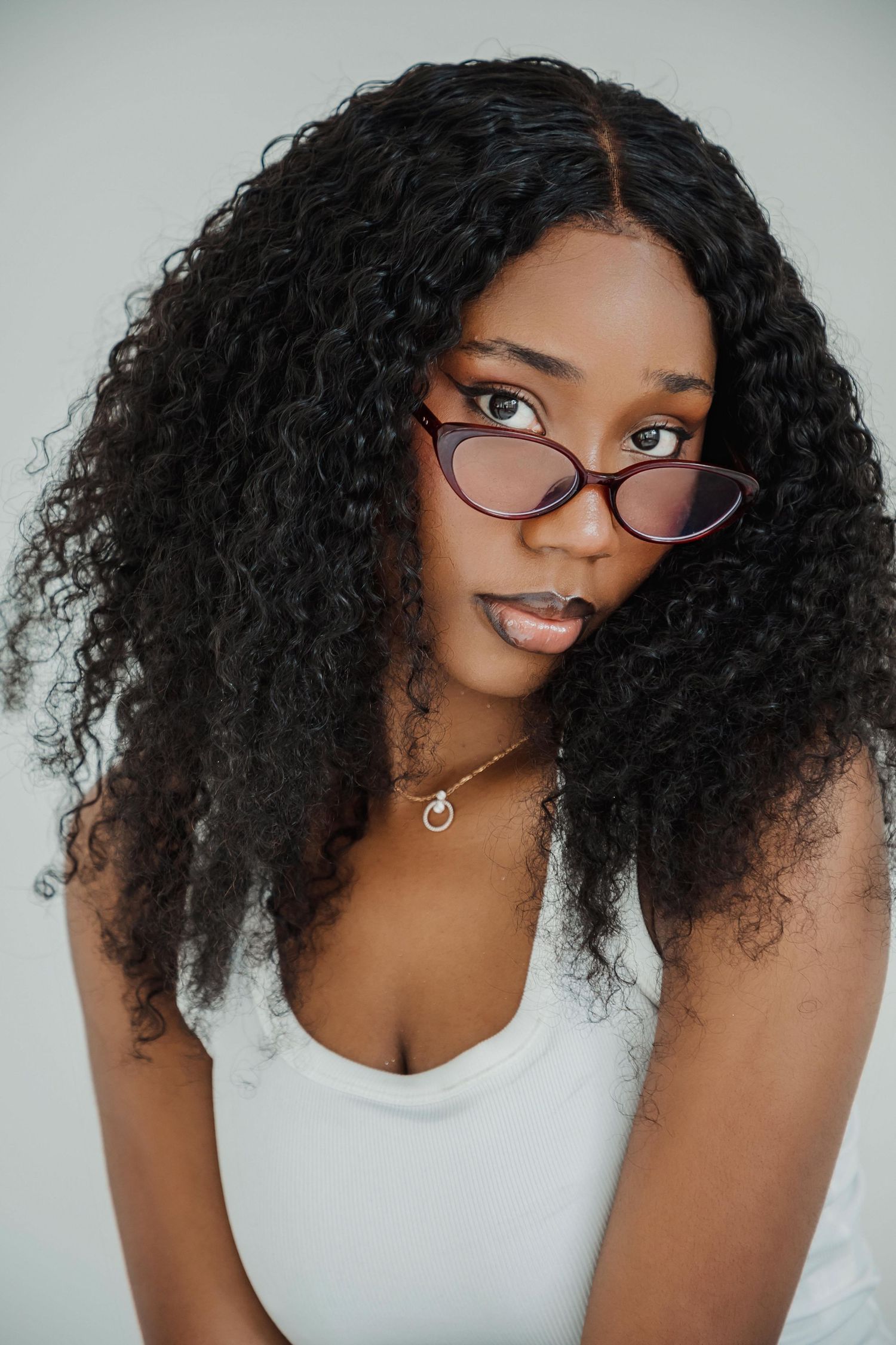 Young Black woman with long curly hair wearing cat-eye glasses and white top, posing against light background.
