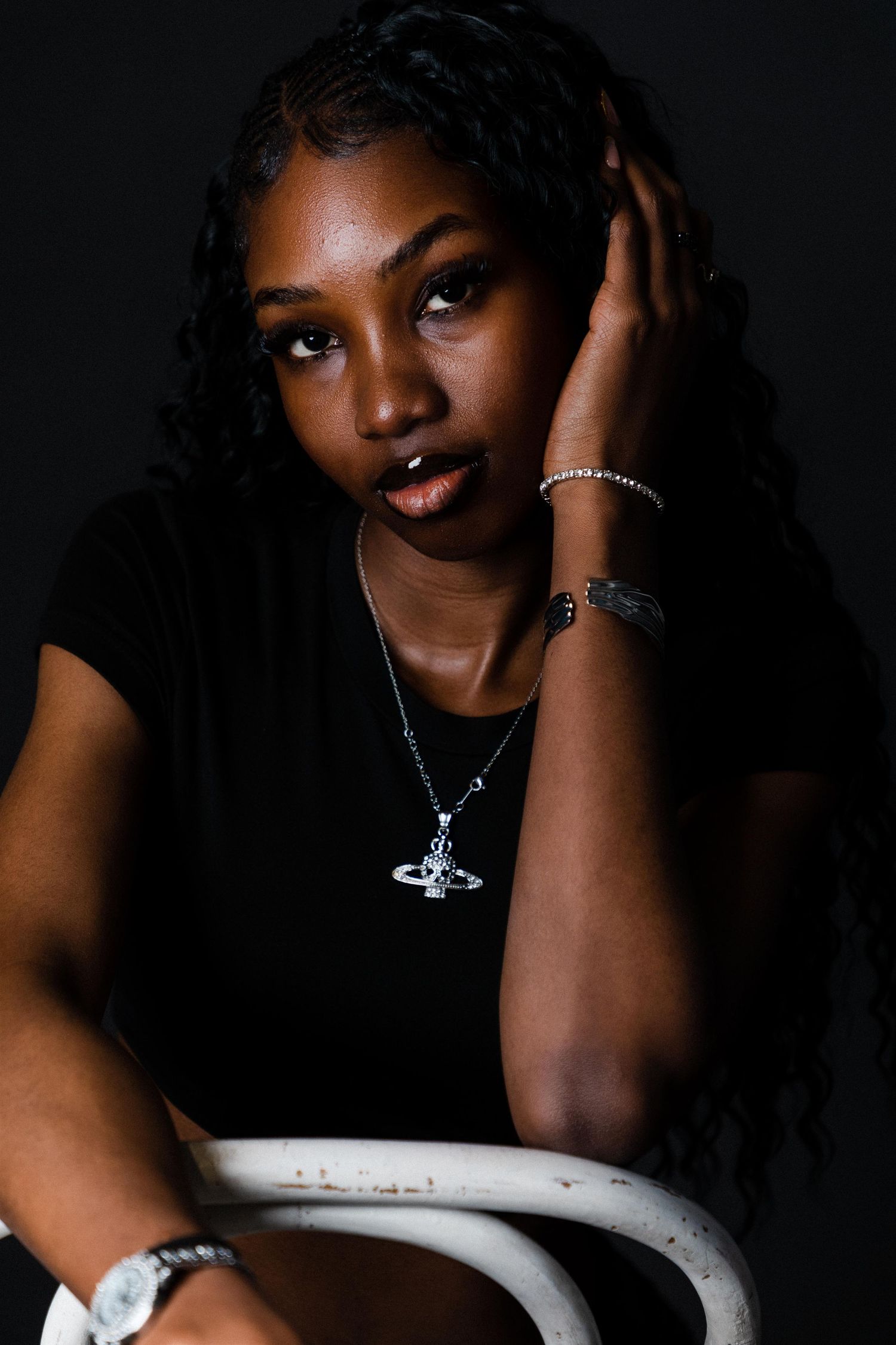 Young Black woman in black tee poses thoughtfully against dark background, wearing Saturn pendant necklace and bracelets.