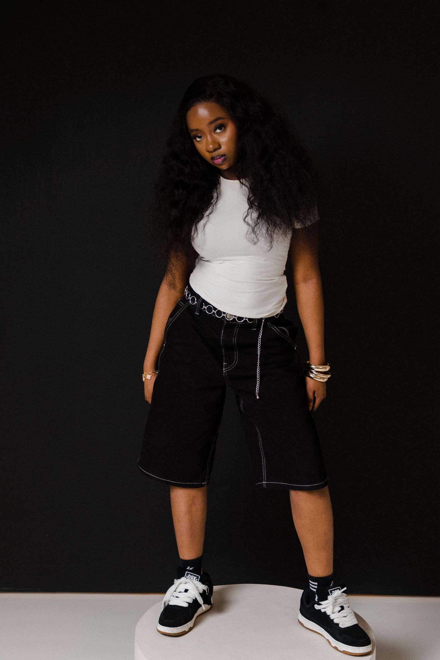 Young Black woman in white crop top, black denim shorts, and sneakers posing against dark background.