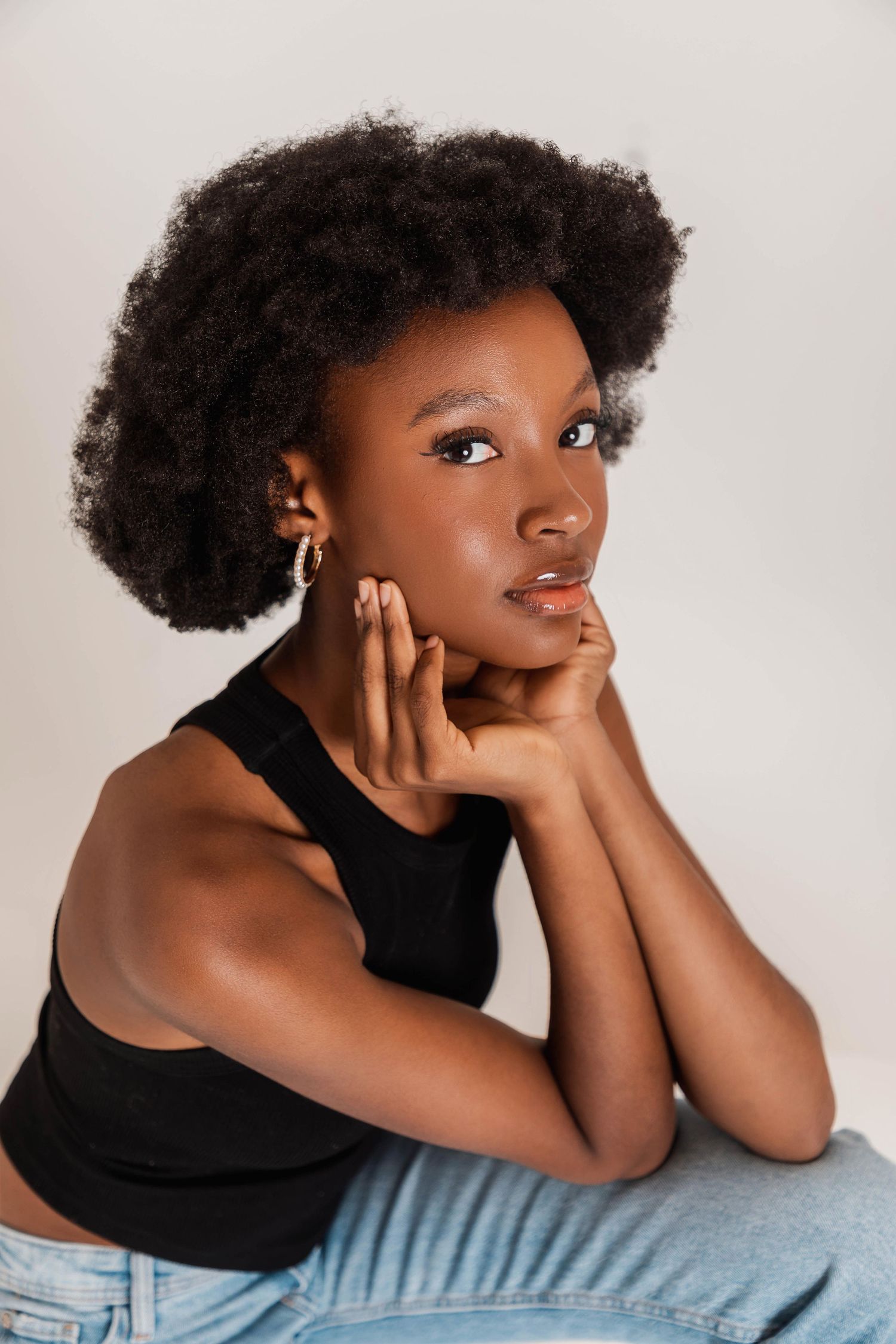 Young Black woman with afro hair wearing black tank top and jeans, posing thoughtfully with hands near face.