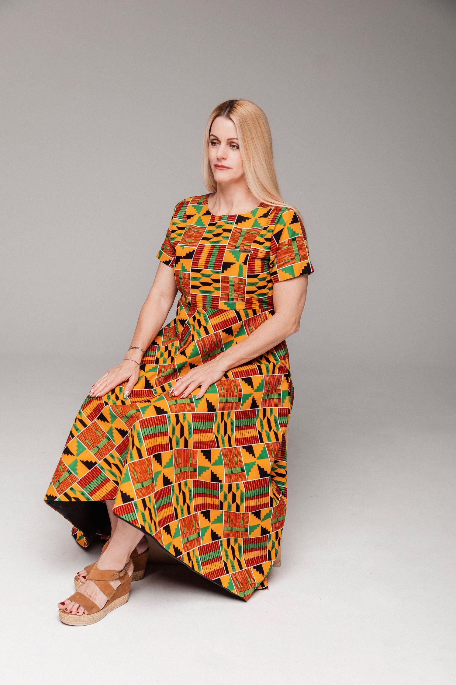 Woman in colorful African kente print maxi dress with short sleeves, seated gracefully on gray background.