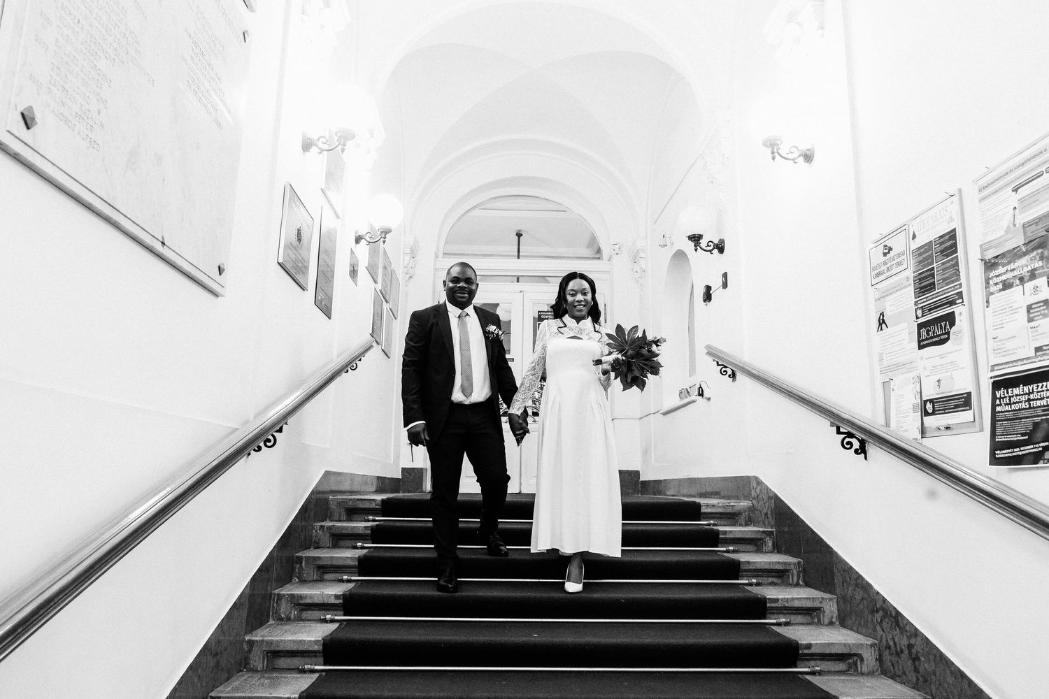 Newlywed couple walks joyfully down staircase in a classic building, bride holding a bouquet.