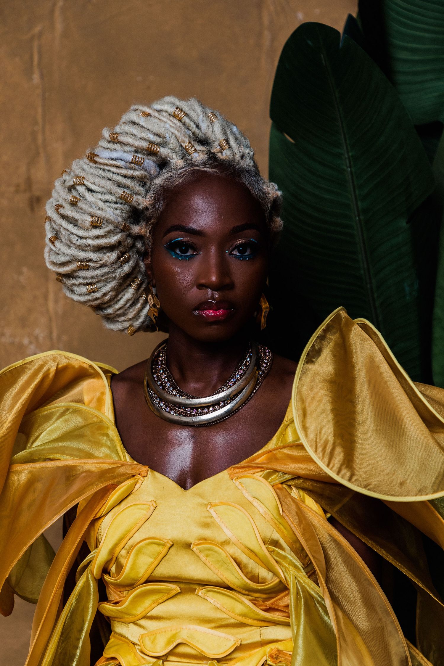 A Black woman in a yellow ruffled gown with silver necklaces, blue eye makeup, and white locs poses near tropical leaves.