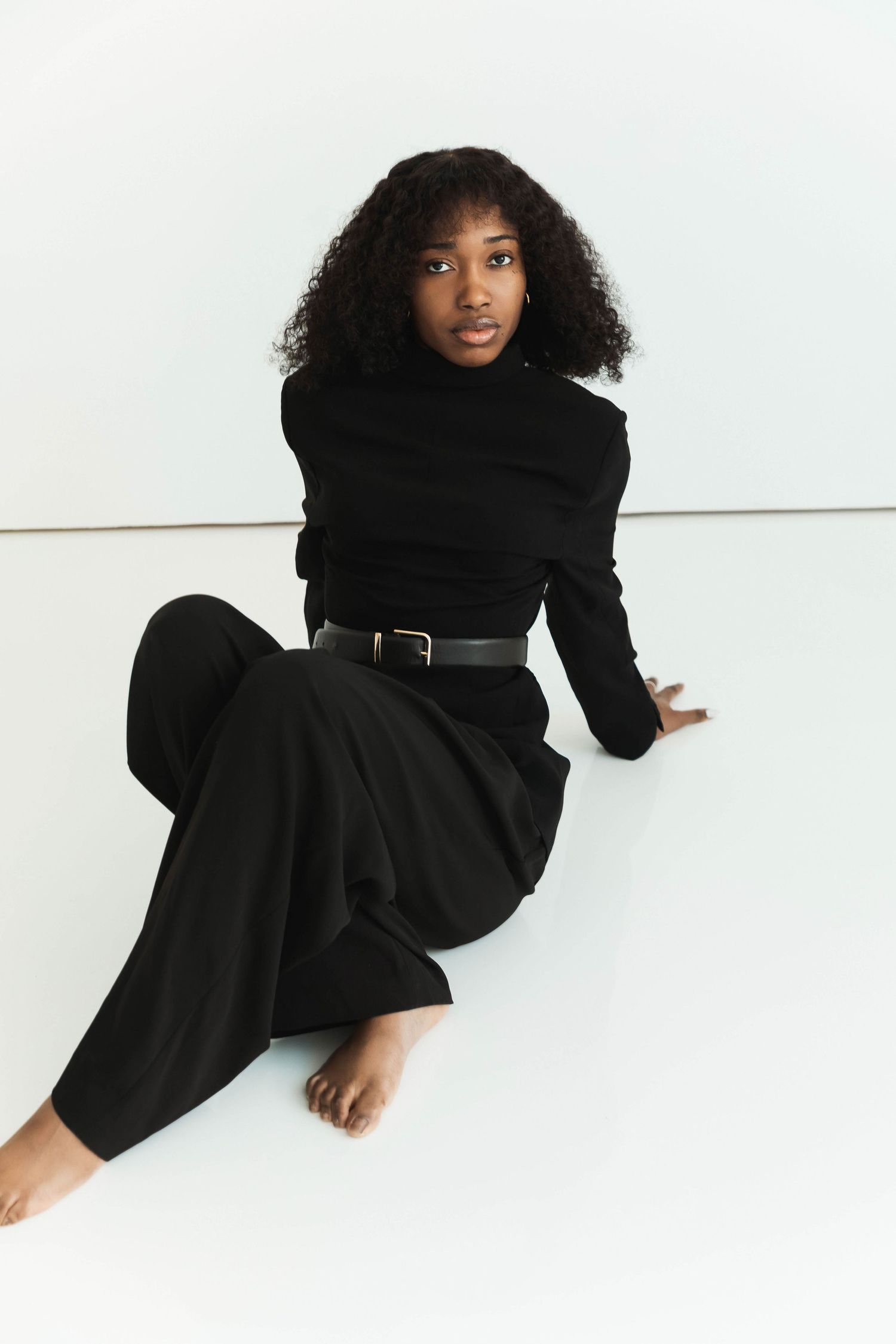 Young woman in all-black outfit with belt sits barefoot on white floor, wearing turtleneck and wide-leg trousers.
