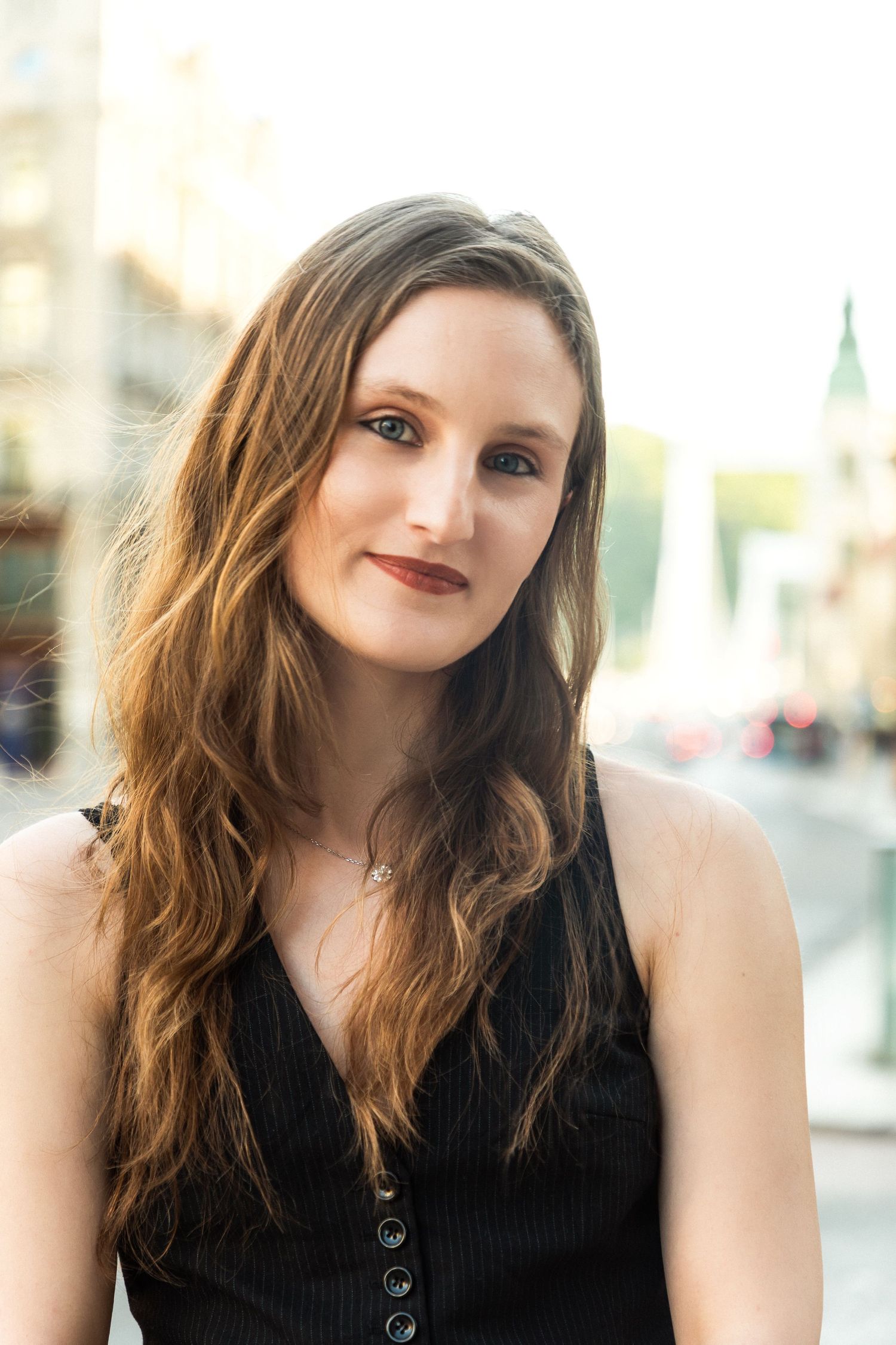 Young woman with long brown hair wearing a black sleeveless top poses outdoors in an urban setting.