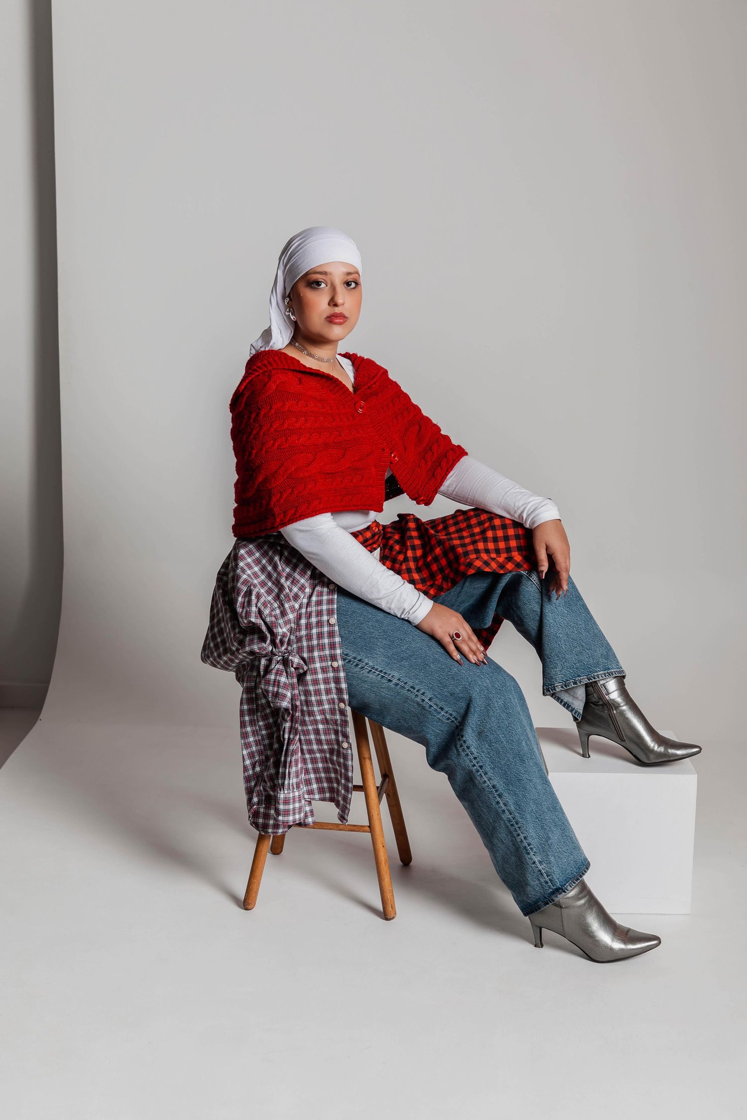 Woman in red knit top, white headscarf, jeans, and silver boots sits on stool in studio setting.
