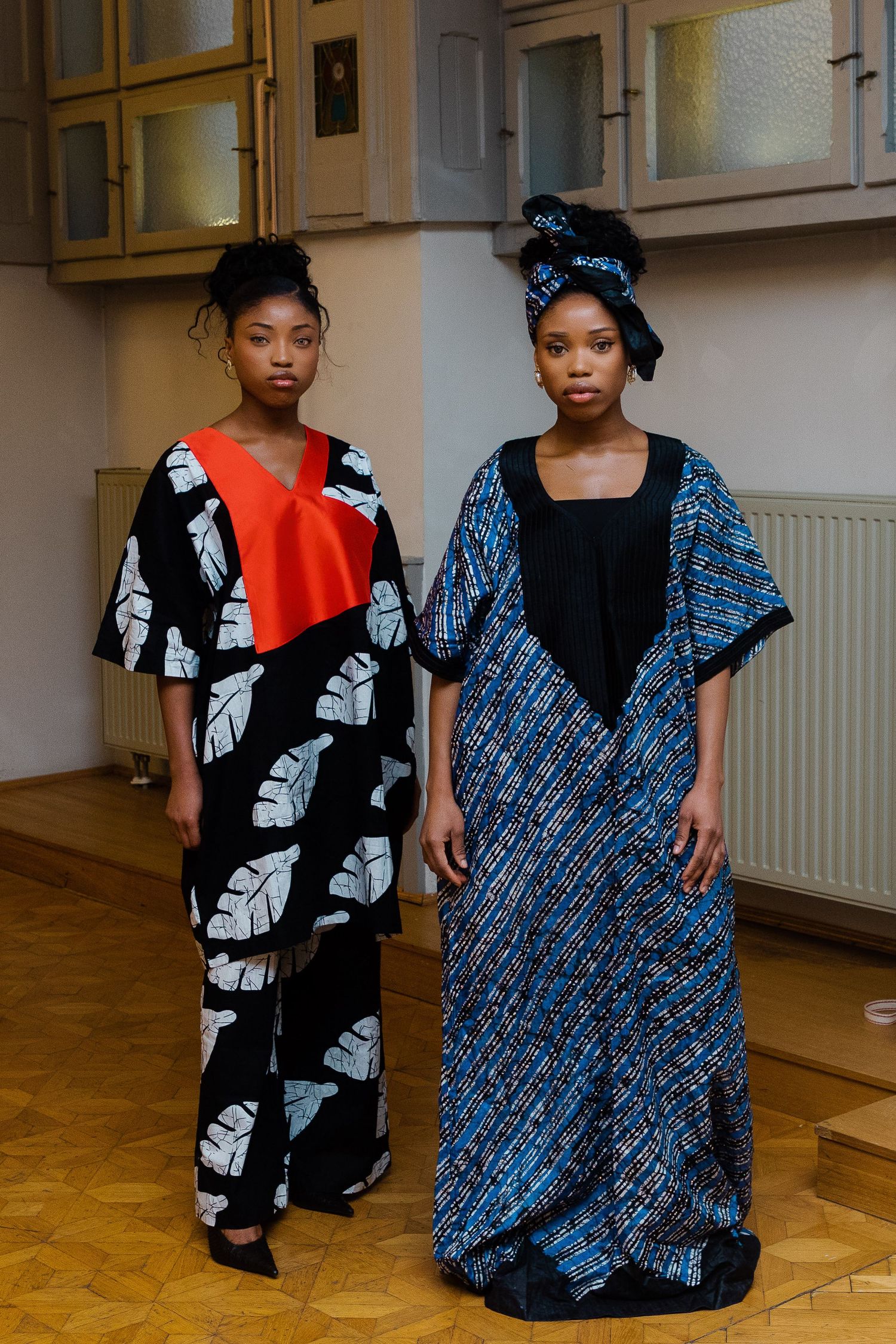 Two women modeling African print fashion designs, featuring bold patterns in black, white, red, and blue traditional fabrics.