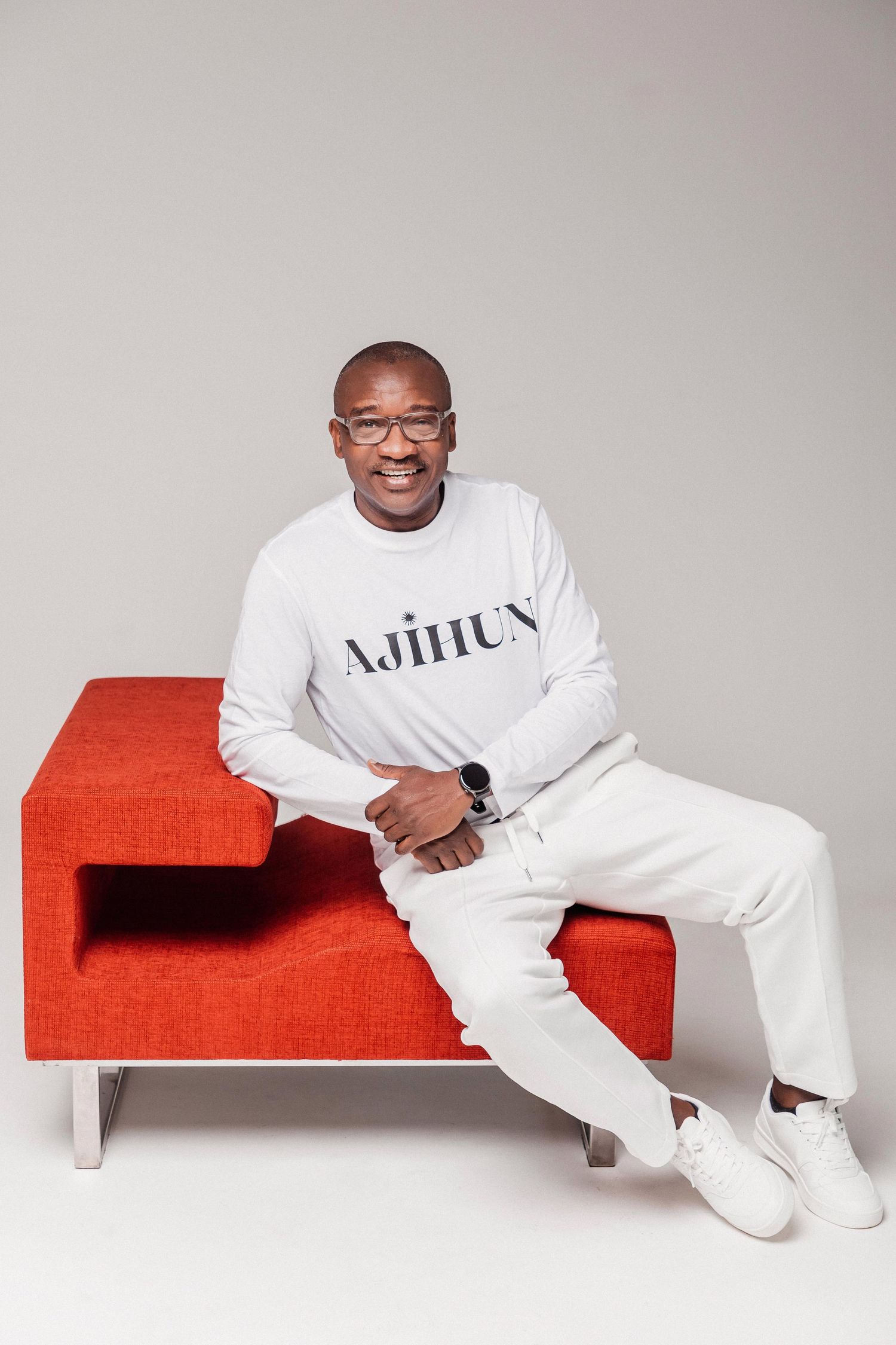 Smiling man in white AJIHUN sweatshirt and pants sitting on a red bench, wearing glasses and a smartwatch.