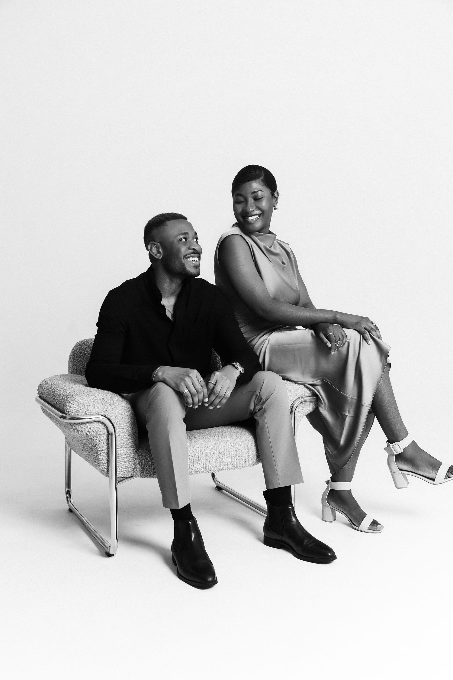 Black and white photo of a smiling couple sitting together on a modern chair in a studio setting.