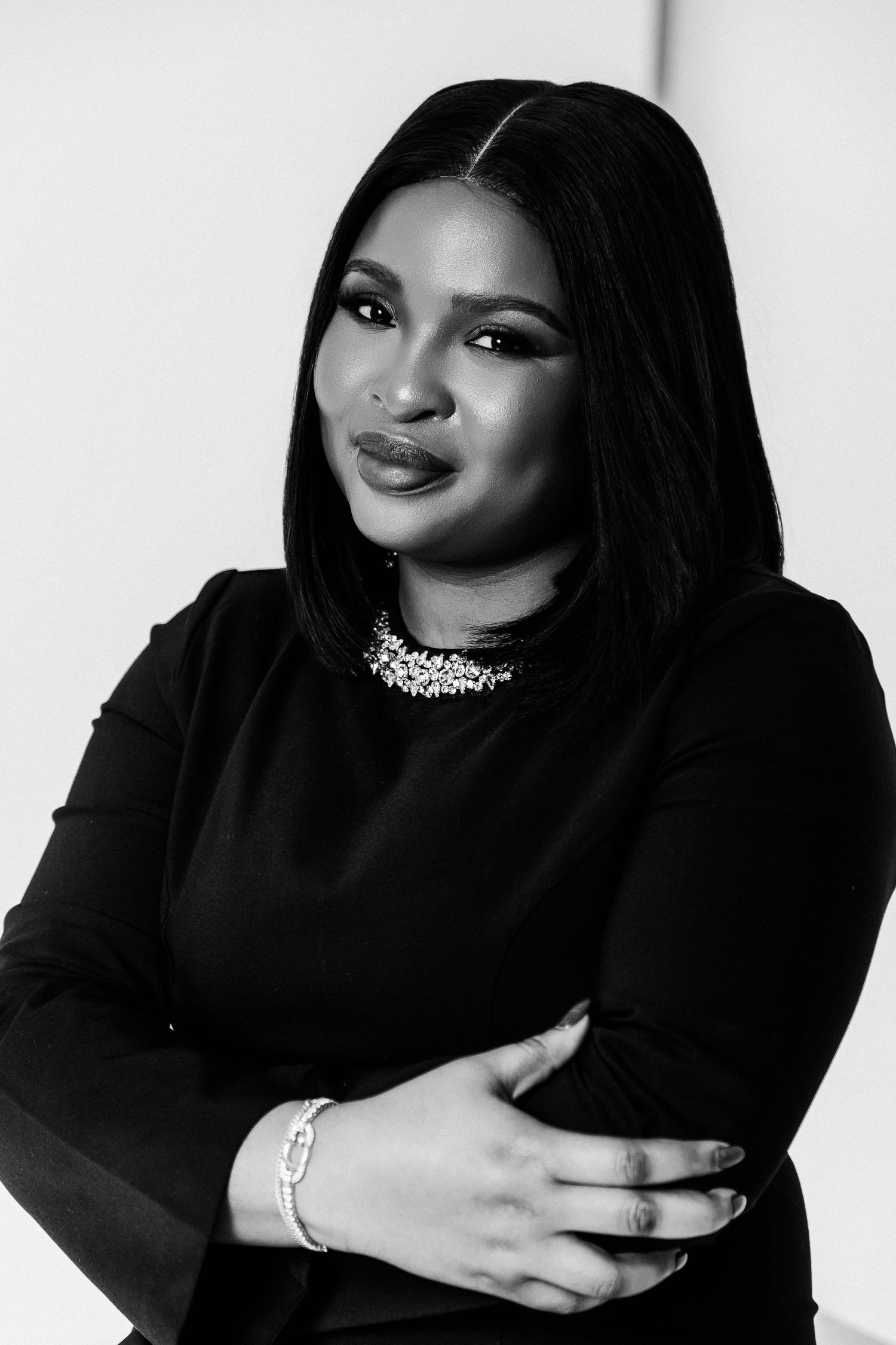 Black and white portrait of a confident woman in black outfit with diamond necklace, arms crossed.