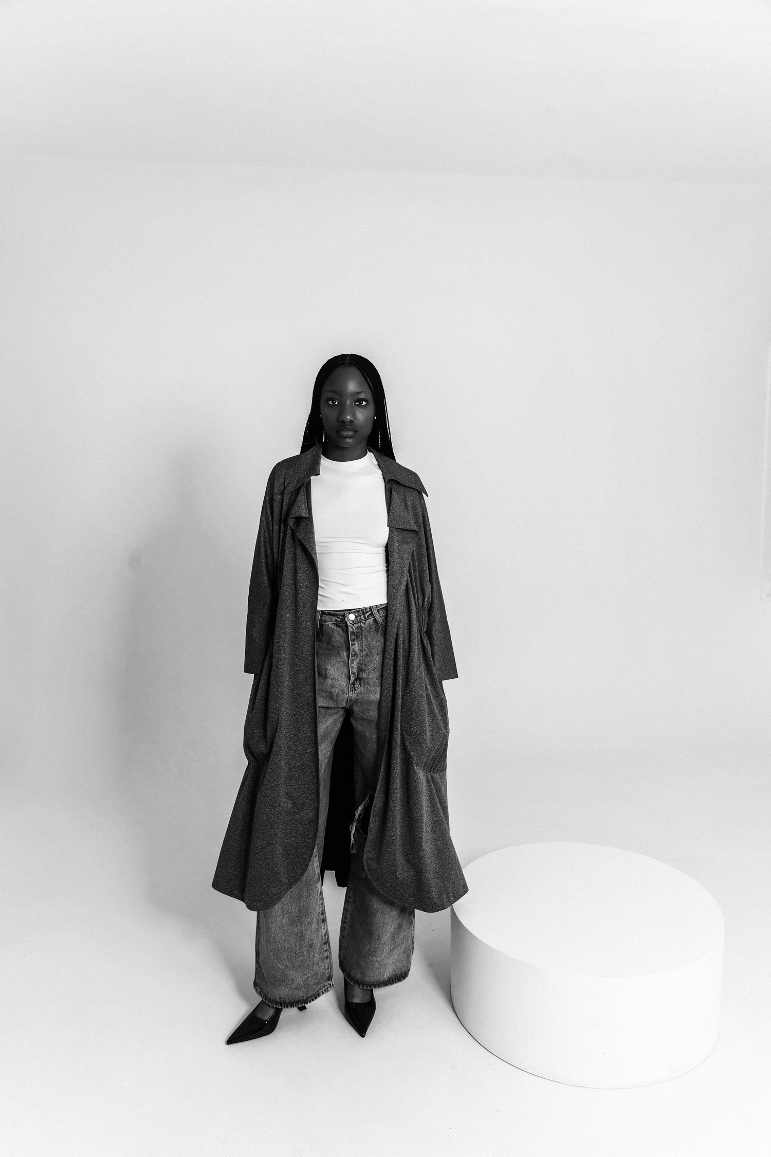 Black and white photo of a woman in a long trench coat, white turtleneck, and wide-leg jeans posing in a studio.